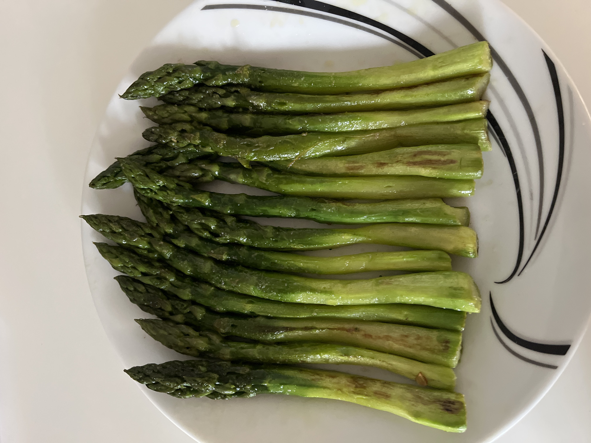 The image presents a close-up, top-down view of a serving of cooked asparagus spears on a white plate. Approximately fifteen to twenty individual asparagus spears are neatly arranged horizontally, filling the central part of the frame.
The asparagus spears are vibrant green, varying slightly in shade from darker tips to lighter, somewhat translucent stems. Many spears exhibit a glossy sheen, indicating they are moist, possibly from steaming, boiling, or being lightly coated in oil. Some of the lower stem sections show subtle brown or reddish-brown discoloration, which can occur during cooking. The tips of the asparagus are firm and intact.
The plate itself is white with a distinctive decorative pattern of several thin, curved black and grey lines appearing on the right side. The background is a plain, light-colored surface, likely a table or countertop. There are no people visible, and no discernible text. The lighting is bright and even, highlighting the textures and colors of the food.
