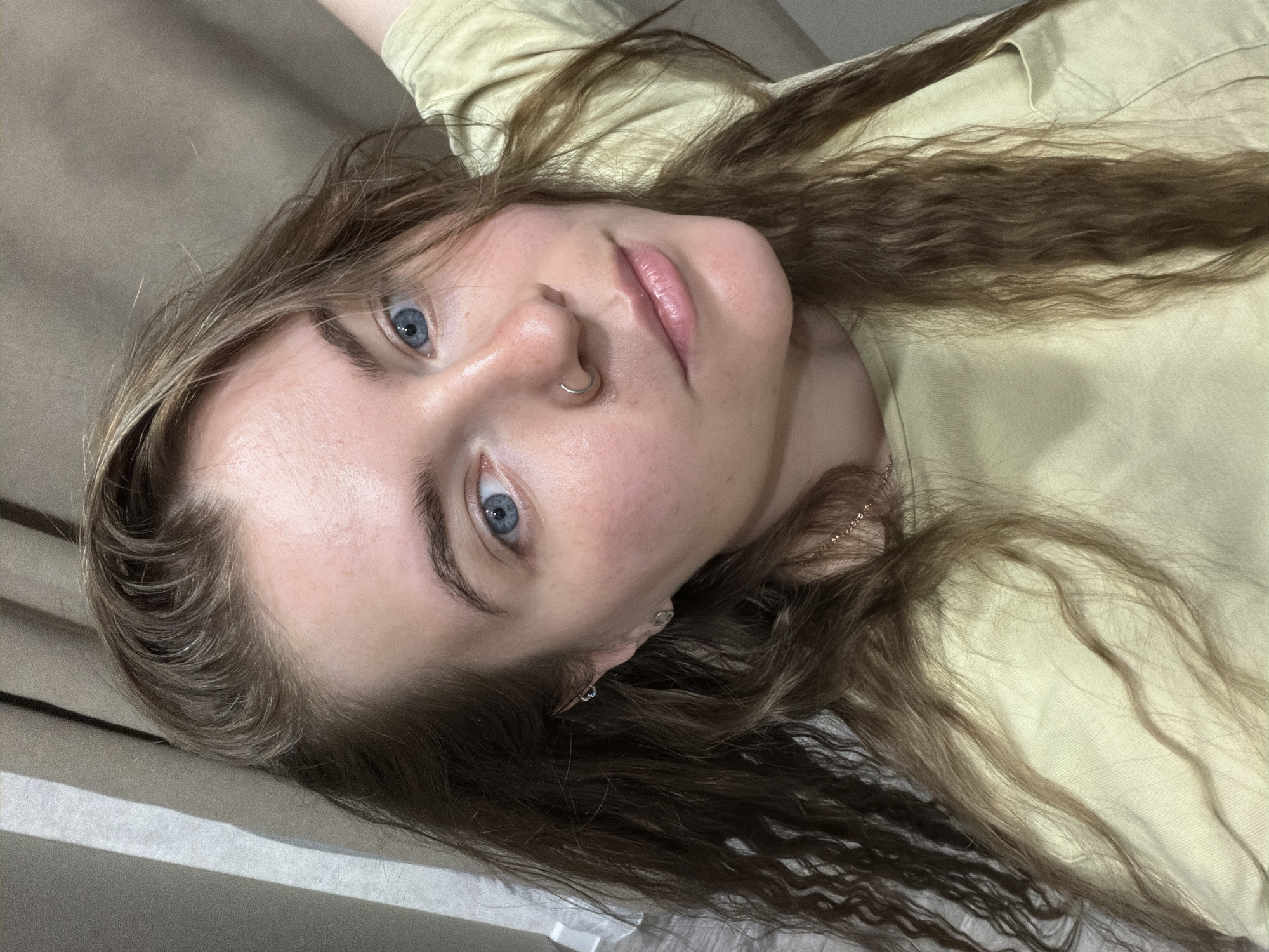 A young woman with long, wavy brown hair and blue eyes is lying on a light-colored surface, possibly a bed or sofa. She has a nose ring and appears to be wearing earrings. The woman is wearing a light yellow t-shirt. The background is blurred, but appears to be a neutral tone, possibly a wall or fabric. The scene is indoors and there is no evidence of the location being in the city of Chaikovsky, Russia. The image is a close-up, focusing on the woman's face.