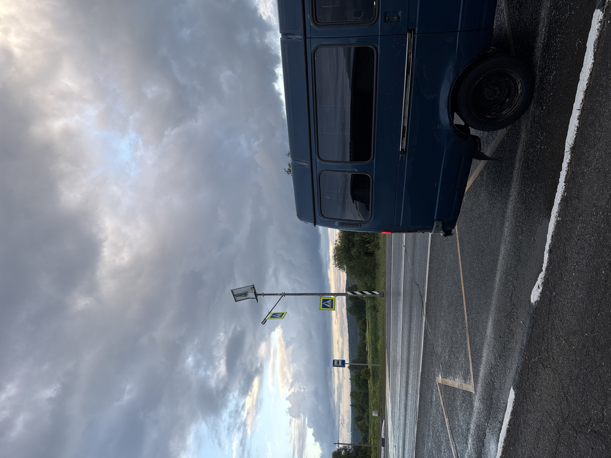 The image depicts a roadside scene in Polozovo, Russia, under a dramatic, overcast sky. A dark blue van is parked on the right side of the frame, its rear end visible. The van has tinted windows and a chrome strip along its side. In the foreground, a wet asphalt road is marked with white and yellow lines, indicating a crosswalk or intersection.
To the left of the van, a tall lamppost stands with two yellow pedestrian crossing signs, one above the other. A blue bus stop sign is also visible in the distance, further left. The background features a lush green landscape of trees and fields, with rolling hills under a vast, cloudy sky. The clouds are a mix of dark grays and lighter whites, with hints of pale blue sky peeking through in the upper left corner. The overall impression is one of a rural or semi-rural location at dusk or during a period of inclement weather. There are no people visible in the image.