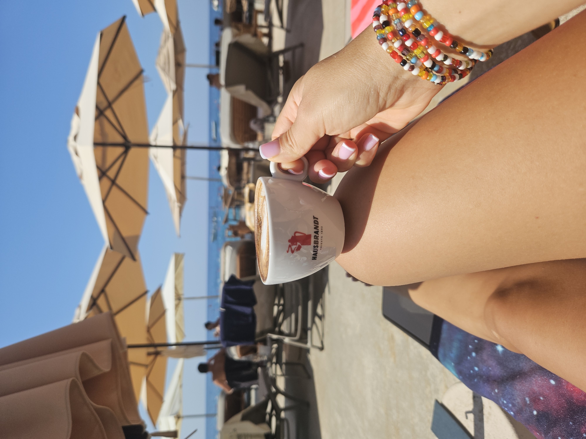 An over-the-shoulder shot shows a person's hand holding a white espresso cup with a Hausbrandt logo. The cup is filled with coffee with latte art. The person is wearing a colorful beaded bracelet on their wrist. The background features a beach with rows of lounge chairs and umbrellas. The ocean and a clear blue sky are visible in the distance. Several people are seen relaxing on the beach.