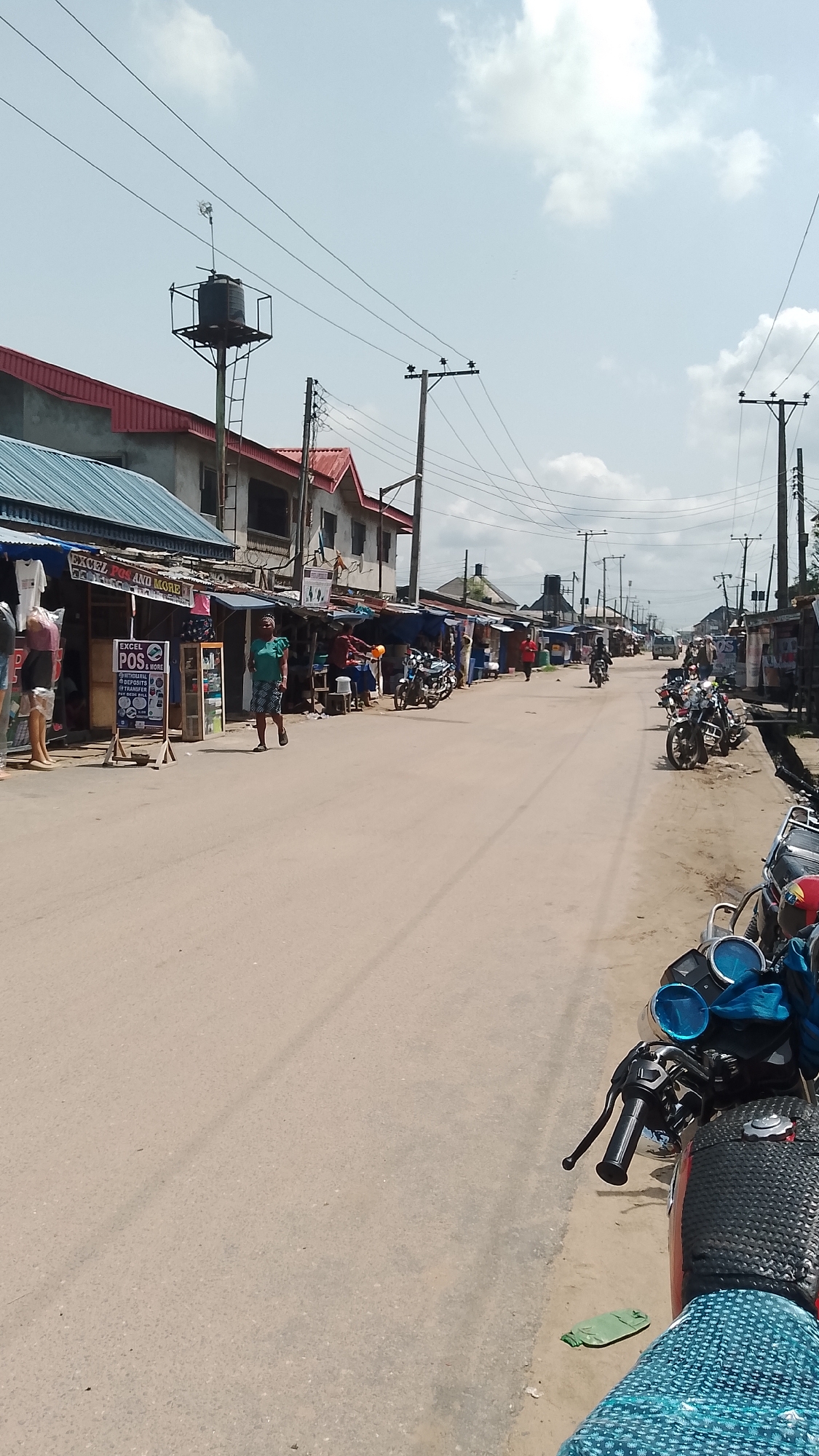 The image depicts a street scene in an urban area of Unknown, Unknown. A paved road stretches into the distance, flanked by buildings and shops on both sides. These structures appear to be a mix of residential and commercial spaces, some with corrugated metal roofs and makeshift awnings.
Several motorcycles are parked along the road, adding to the bustling atmosphere. A few individuals are visible: one person walks along the road, while others appear to be tending to shops or engaging in activities along the street.
Overhead, utility poles and wires crisscross the sky. A water tank on a metal structure is seen atop one of the buildings. The sky is partly cloudy.