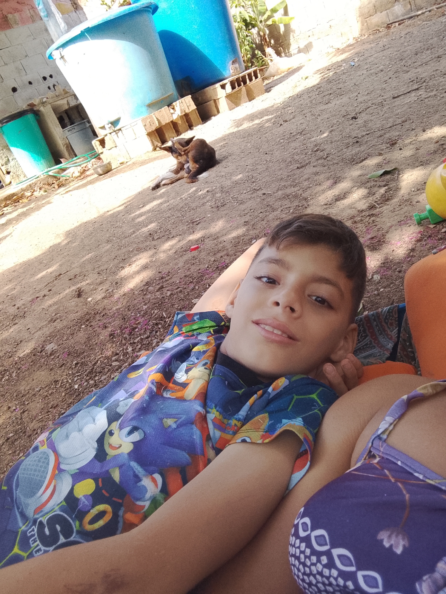 A young adolescent boy is lying down, appearing to rest his head on the lap or chest of an unseen individual. He is wearing a blue shirt with Sonic the Hedgehog characters. The unseen person is wearing a purple top with a white pattern. In the background, there is a brown dog lying on the ground, next to some blue water tanks supported by cinder blocks. The ground is dirt. This photo was taken in Altagracia, Venezuela.