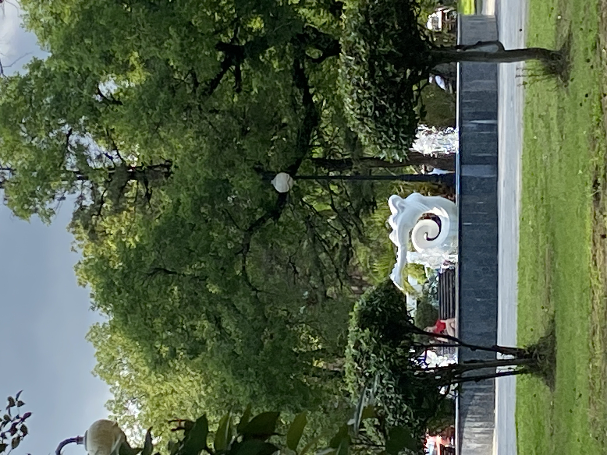 In Sokhumi, Abkhazia, a public park features a circular fountain with a modern sculpture of a white spiral in its center. Water jets erupt from the fountain's edges. Lush green trees with thick foliage form a dense canopy overhead, casting shadows on the grassy ground. A path runs in front of the fountain, and on the path, to the right of the fountain, a person in a red top and dark pants sits on a bench. The park also includes manicured bushes in the foreground, one to the left and one to the right of the frame. A lamppost with a rounded white lamp stands behind the fountain. The sky above is a pale blue with some clouds.