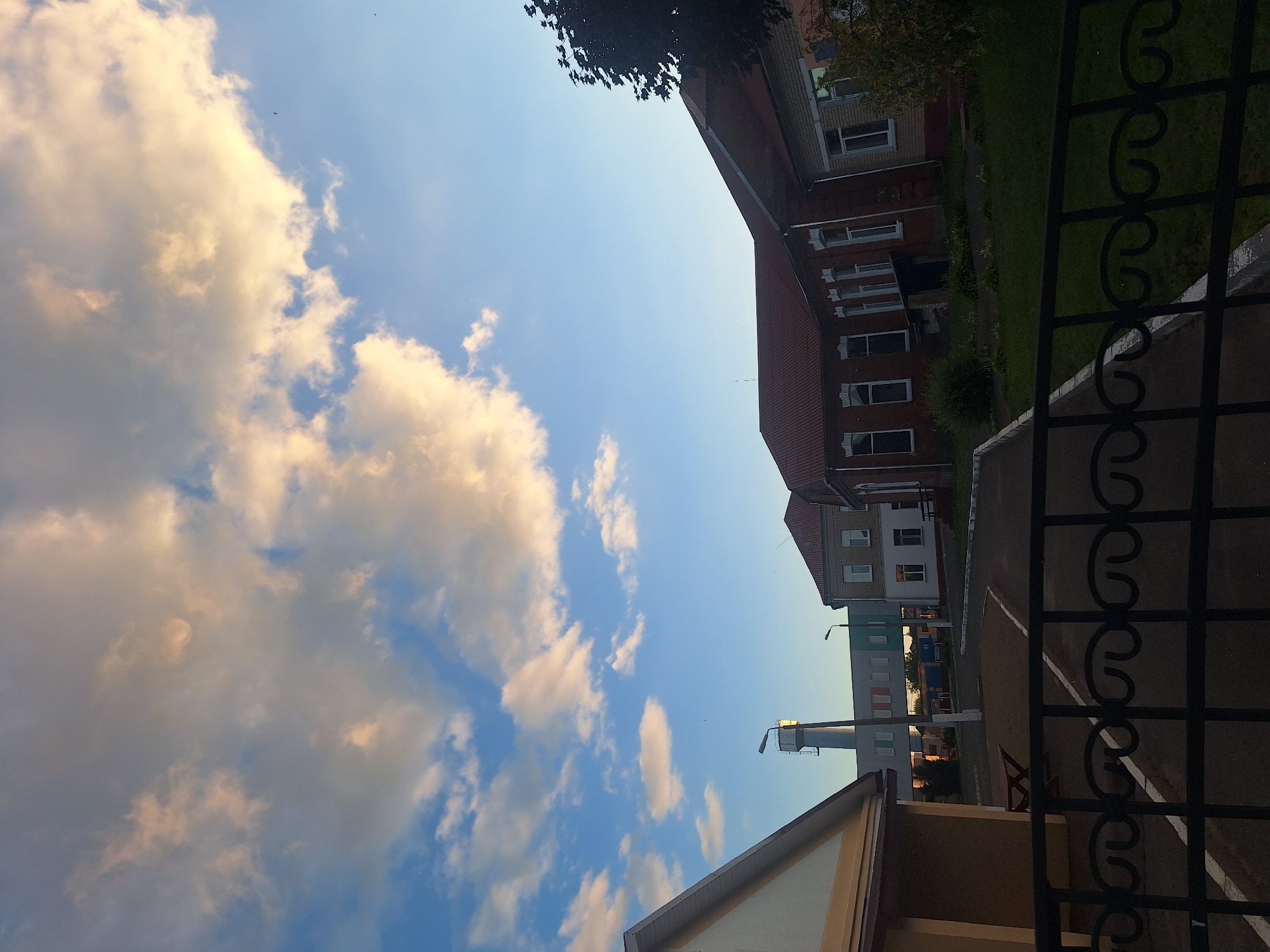 The image captures a view of buildings and a sky filled with clouds. The architecture suggests a public or institutional setting, possibly a school or administrative complex. The buildings appear to be made of brick and have multiple windows. The sky is blue with scattered clouds, some of which are illuminated by the sun, indicating it might be late afternoon or early morning. There is a metal fence in the foreground, suggesting the image was taken from behind a boundary. The overall impression is one of a serene, daytime scene in a somewhat utilitarian environment, possibly in the Капыльскі сельскі Савет region of Belarus.