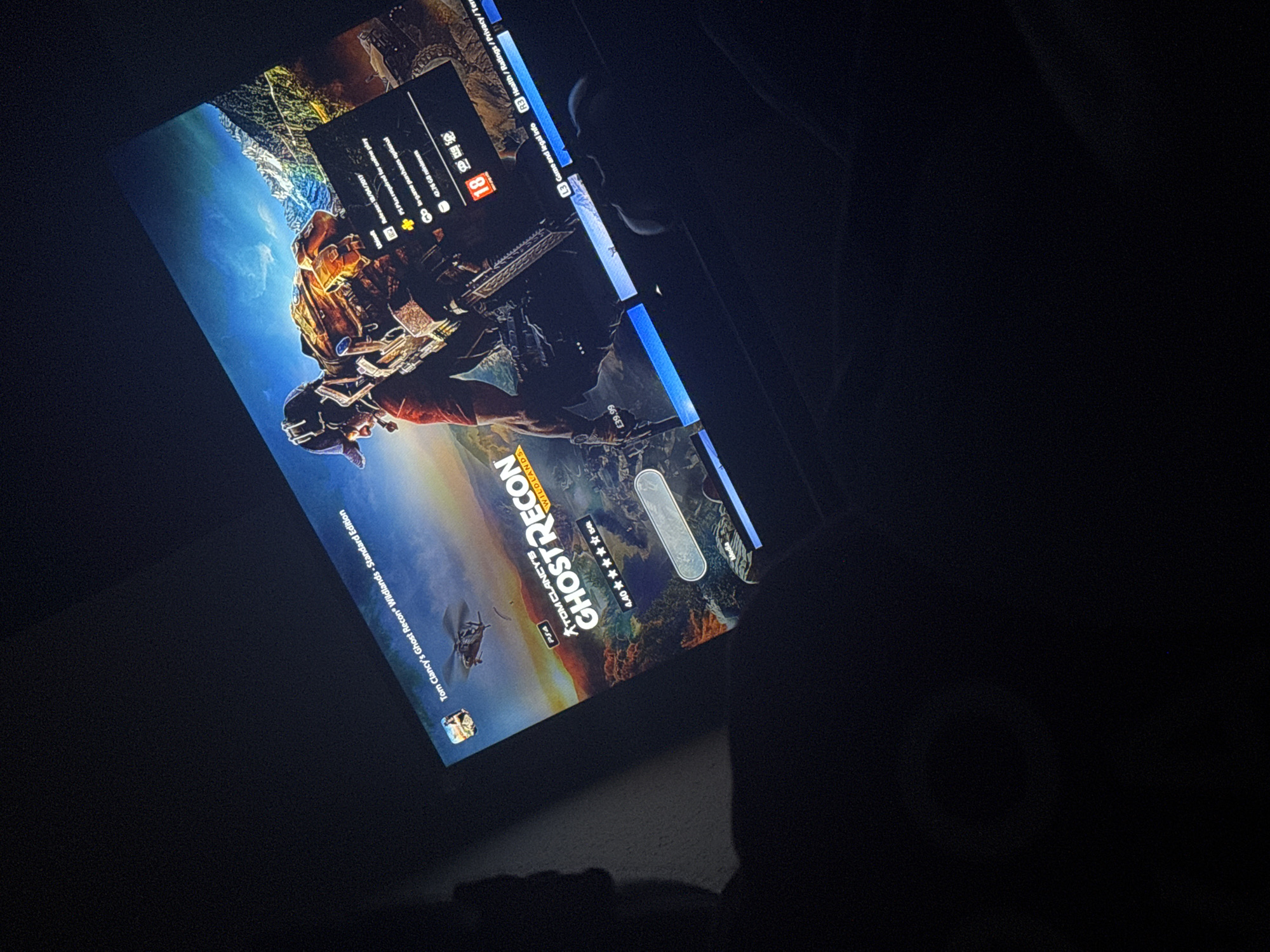 Here's a forensic description of the image:
The image shows a dark, low-lit environment. The primary subject is a screen displaying the game "Tom Clancy's Ghost Recon Wildlands - Standard Edition" on a PlayStation platform. The game's artwork features a soldier figure and a landscape with a blue sky. Text on the screen includes "Ghost Recon Wildlands," "4.40 stars" rating, a price of "£39.99," and information about storage size (42.76 GB minimum). The "PSA" and "Ubisoft" logos are visible. There is a rating label "18" on the screen, along with text related to in-game content and legal information. Parts of the surroundings are visible, including a dark surface and what appears to be a person's form partially visible in the darkness, suggesting the photo was taken indoors. There is no indication of the specific location within Welshpool, United Kingdom, nor any indication it is even located there.