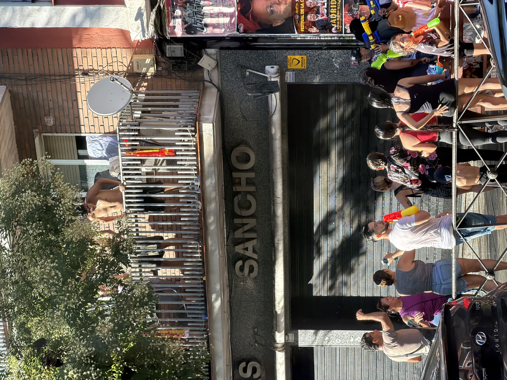 The image depicts a lively street scene, likely during a summer festival or celebration in Madrid, Spain. In the foreground, a group of people are gathered, many holding water guns, suggesting a water fight. Some individuals are smiling and engaged in the activity, while others are observing. Behind them, a closed storefront with the prominent sign "SANCHO" is visible. Above the storefront, on a balcony, several individuals are present, with one man shirtless and leaning over the railing. The environment appears to be a typical urban setting with buildings, a balcony with railings, and trees providing shade. The overall atmosphere is one of casual fun and social interaction, characteristic of a public gathering in a city.