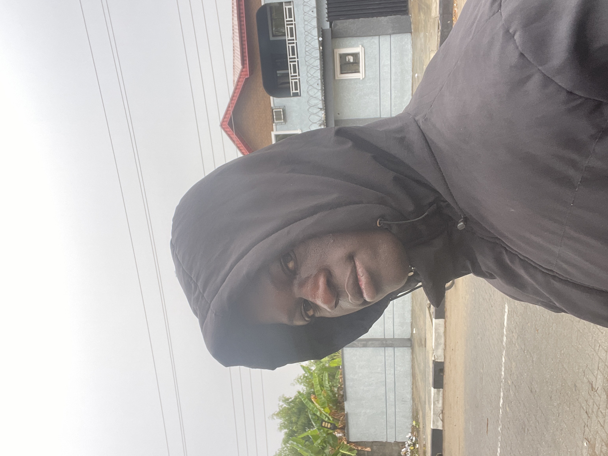 A person wearing a black hooded jacket stands outdoors on a pavement. A two-story building in the background features barred windows, light-colored walls, and a red roof. Power lines are visible in the sky. Vegetation, including banana plants, grows near the base of a concrete fence. The location is in Badagry, Nigeria.