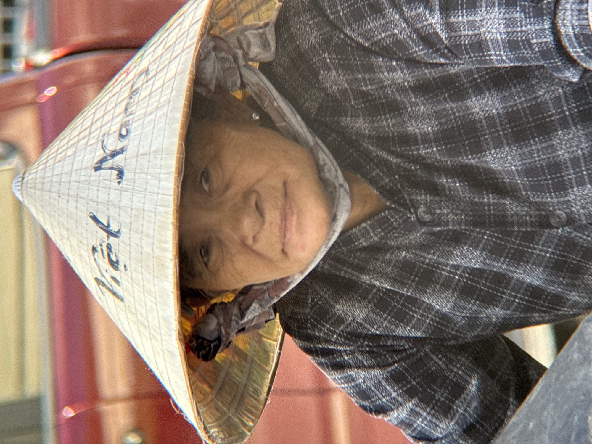 A close-up portrait shows an elderly Vietnamese woman wearing a conical hat inscribed with the words "Việt Nam." She has a patterned scarf draped around her neck and chest and is wearing a plaid shirt. The background is blurred, but a red vehicle is visible. The image appears to have been taken outdoors in a public area, possibly in Nha Trang, Vietnam, given the inscription on the hat.