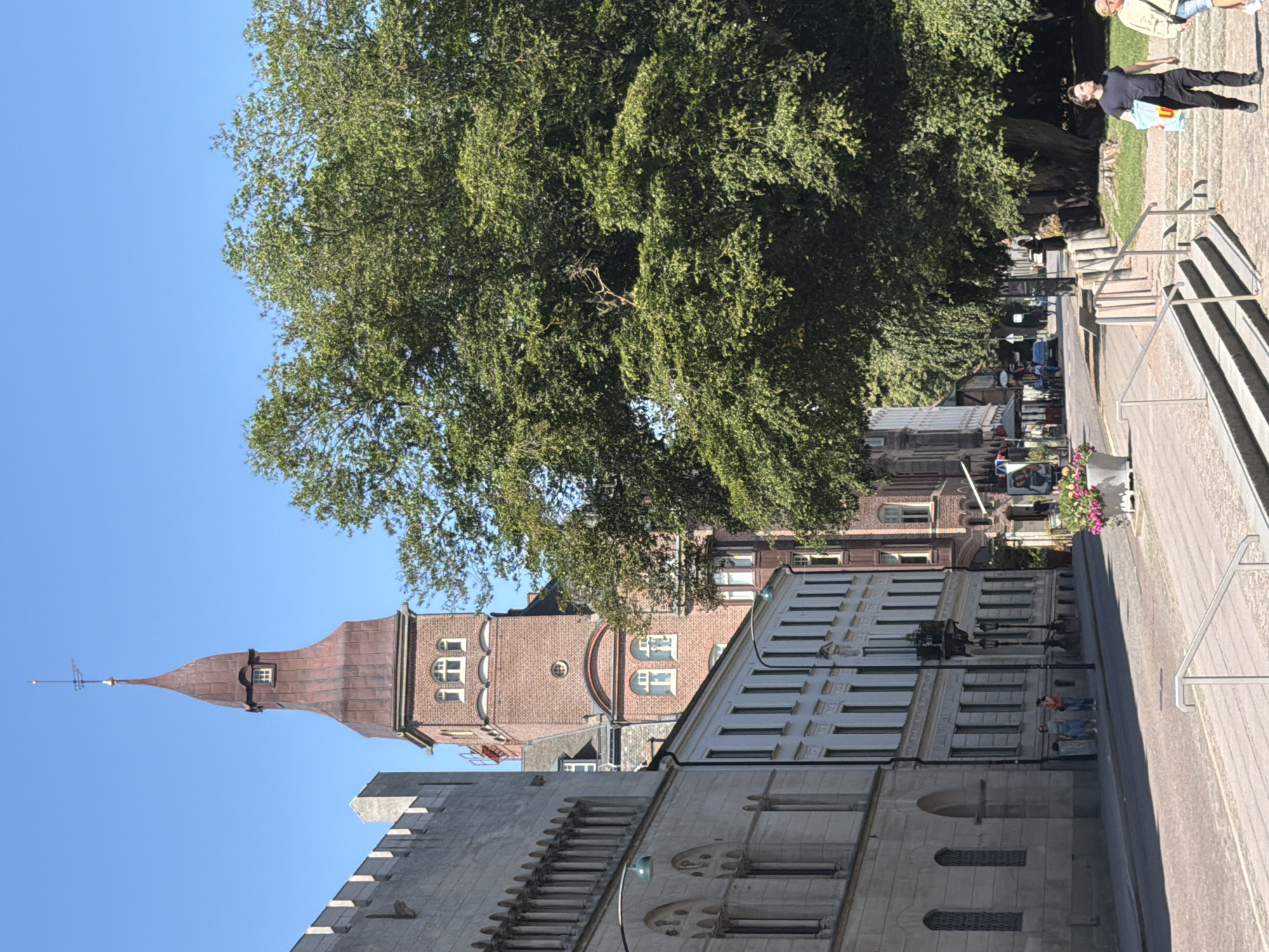The image depicts a sunlit street scene in Lund, Sweden. In the foreground, a paved walkway with metal railings leads down a set of stone steps. Two individuals are descending the steps; one, a man wearing a dark t-shirt and black trousers, carries a blue shopping bag. Beside him, a person in a light-colored top and shorts walks. To the right, a large, leafy tree dominates the scene. Across the street, a row of buildings lines the sidewalk, with a tall, reddish-brown brick tower featuring a copper-colored spire and a weather vane prominently visible. The tower has arched windows and decorative brickwork. Further down the street, other buildings and trees are visible, and several people can be seen walking or standing on the sidewalk. A car is also present on the street. The sky is clear and blue. The architecture suggests a historical European city. The tower is likely the Old University Building (Universitetsbiblioteket) due to its distinctive architecture and location in Lund.