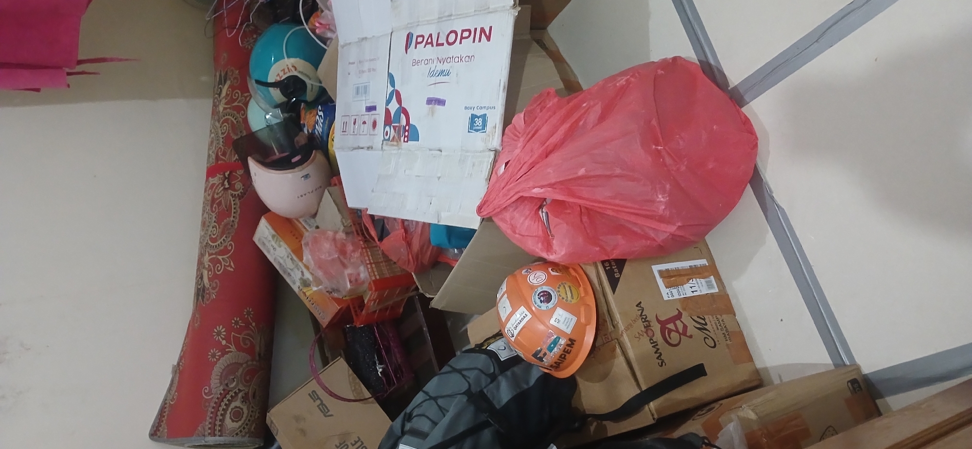 A cluttered indoor scene shows a floor and wall, with various objects piled around. On the floor, there is a large, rolled-up red rug with a floral pattern. To the right of the rug are two motorcycle helmets, one pink and one turquoise. Several cardboard boxes are stacked haphazardly. One box has "ASUS" printed on it. A black and grey backpack is visible. An orange hard hat covered in stickers sits on top of a brown cardboard box. A large red plastic bag is prominent in the foreground, and another red bag is visible further back. A pink towel or fabric hangs from the upper right of the frame. The scene suggests a storage area or a room undergoing tidying or unpacking. The location is identified as Tanjung Balai Karimun, Indonesia.