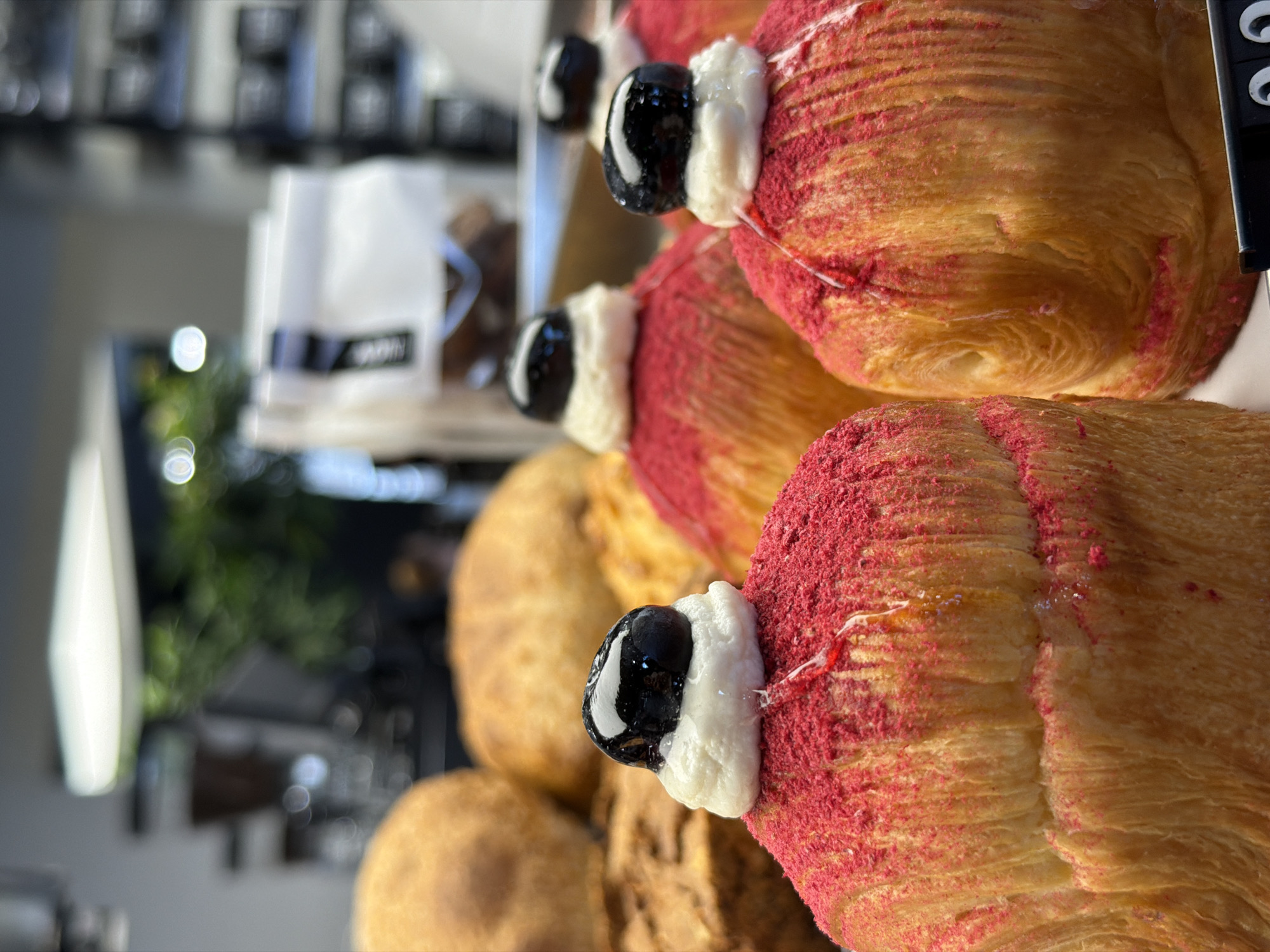 The image showcases a close-up view of several pastries, likely on display in a bakery or cafe. The pastries are golden-brown, flaky, and appear to be a variation of croissants or Danish pastries. The top of the pastries are covered with a red, powdery substance, possibly a berry-flavored topping. Each pastry is adorned with a small, black, shiny, spherical object (possibly a glazed fruit or chocolate ball) held in place by a dollop of white frosting or cream.
In the background, out of focus, there is a display case with other baked goods and a bag with a logo on it. Light fixtures and foliage are visible, suggesting an interior location.
There is no confirmed evidence from this image to suggest it is in Izhevsk, Russia. Without more information, I can only describe the image based on its visual content.