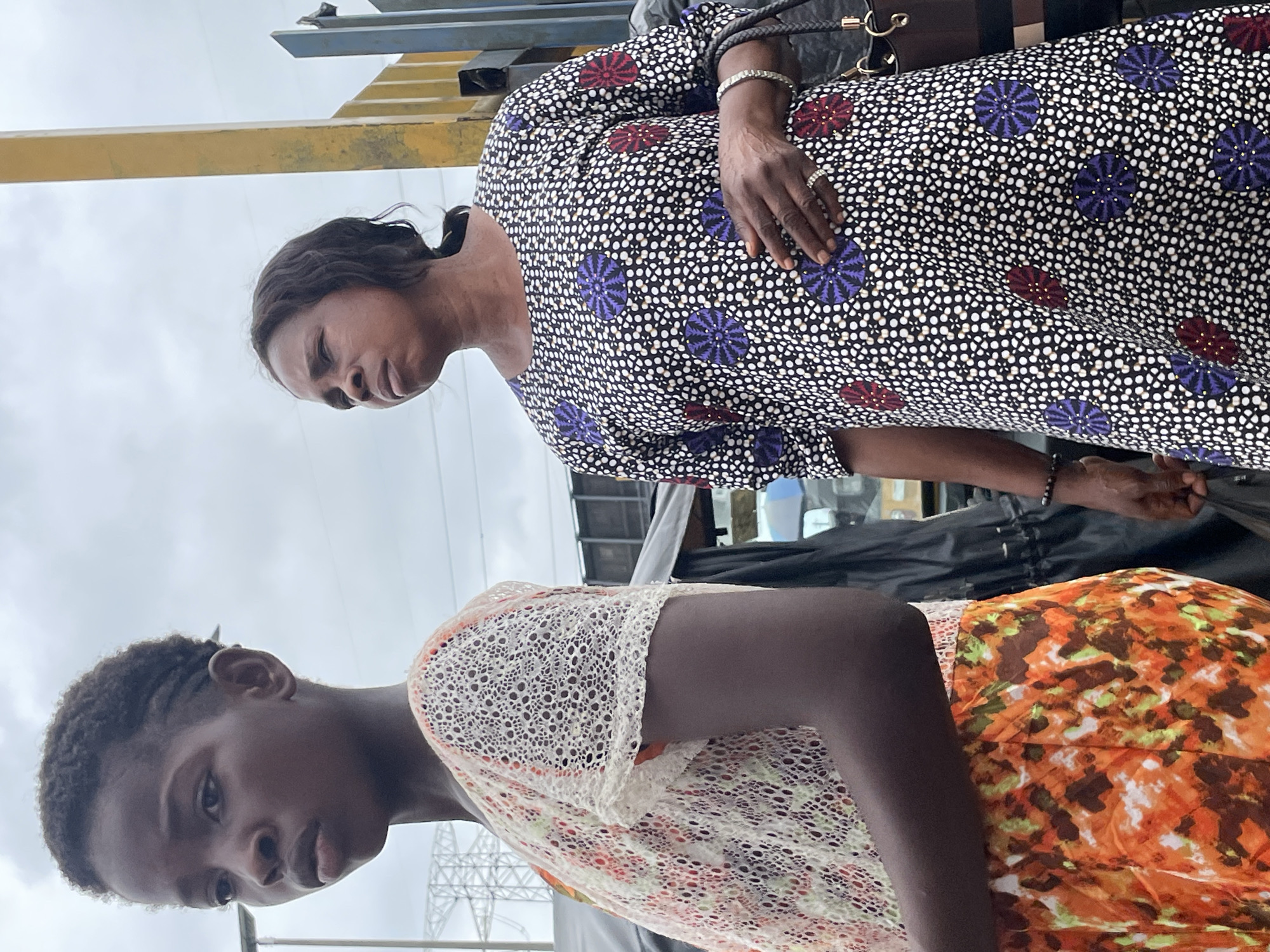 The image shows two people, a younger person on the left and an adult on the right, outdoors in what appears to be a market or street setting in Lekki, Nigeria. The younger person is in the foreground, angled to the left, with dark skin and short, curly hair styled with some braids. They are wearing a white, open-weave top with a lace-like pattern and an orange and green floral print skirt. The adult, positioned behind and to the right of the younger person, has dark skin and dark hair pulled back. They are wearing a black dress with a pattern of white dots and circular floral motifs in purple and red. The adult is holding a dark handbag and wearing a bracelet and a ring. In the background, a yellow metal pole is visible, along with some structures that suggest urban infrastructure, such as power lines and buildings. The sky is overcast.