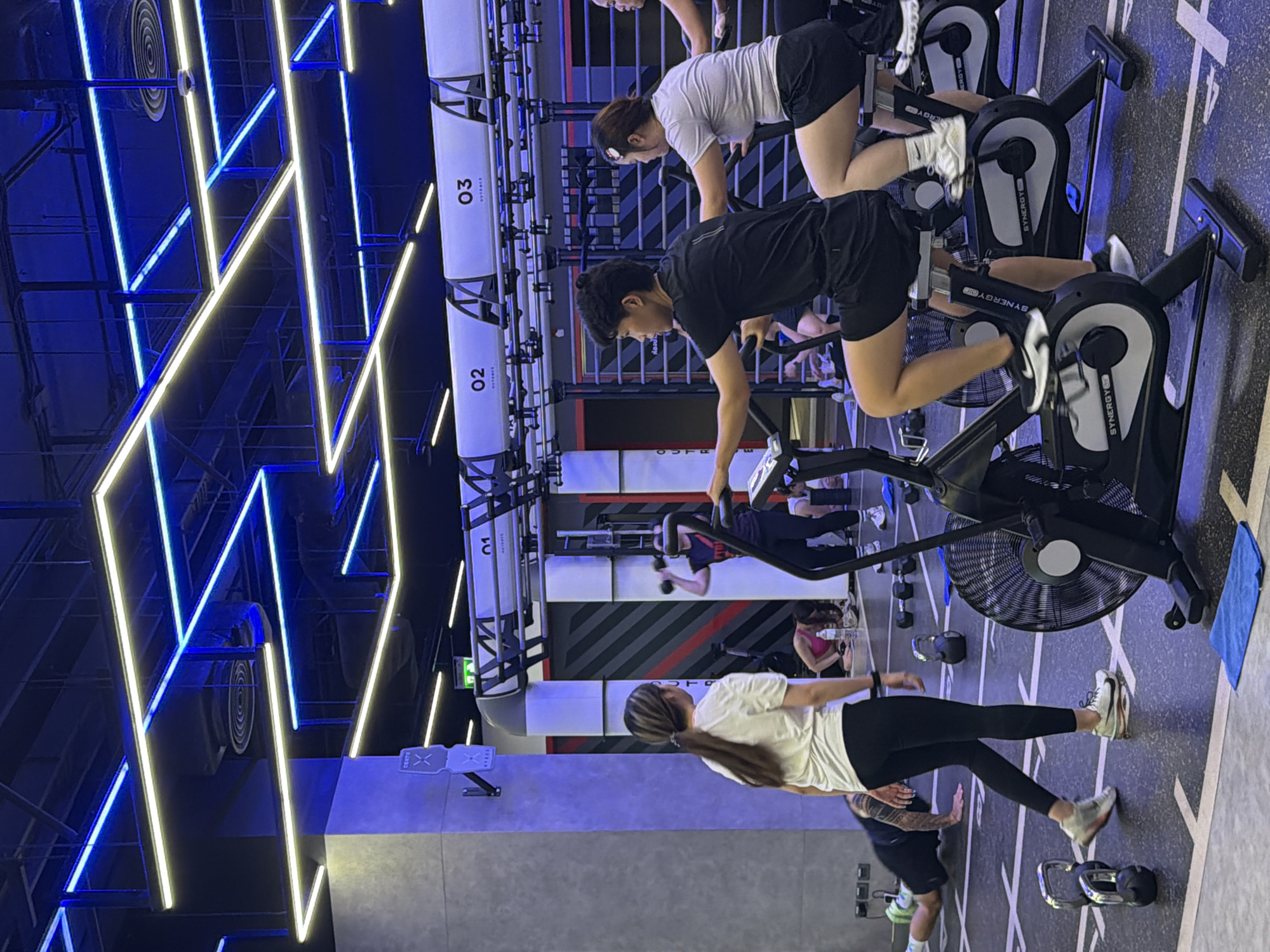In a modern fitness facility, possibly located in Nong Pa Khrang, Thailand, several individuals are engaged in various exercises. In the foreground, two people are seated on stationary bikes, with the person on the left wearing a black t-shirt and shorts, and the person on the right wearing a grey t-shirt and black shorts. To the left of the bikes, a woman in a white t-shirt and black leggings walks away from the camera. Further in the background, another woman is performing a dumbbell exercise overhead. On the floor, a person is in a plank position, and several dumbbells and kettlebells are scattered. The environment is characterized by a dark ceiling with intricate blue and yellow neon lighting, and a grey wall with vertical red and white striped panels. The floor is a dark mat marked with white lines.