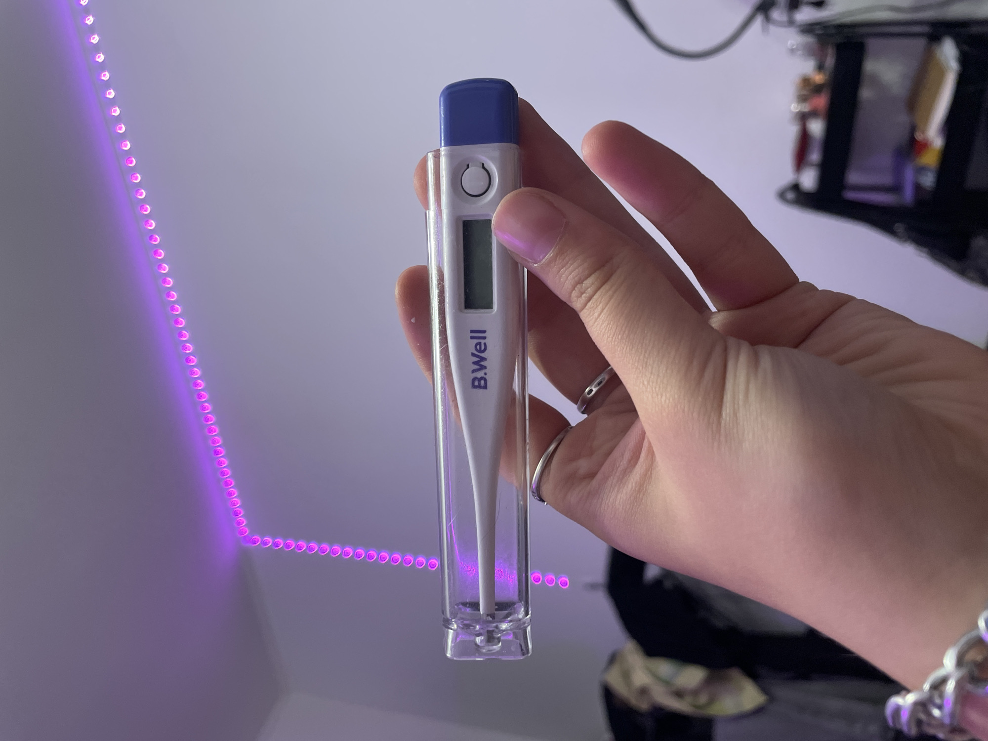 A person's hand is holding a B.Well digital thermometer in a clear plastic case. The thermometer has a white body with a blue tip and a digital display. A purple LED light strip is visible in the background, running along a wall and ceiling. The scene appears to be indoors, possibly a bedroom, with some out-of-focus objects in the background suggesting a domestic setting. The location is Vaganovo, Russia.