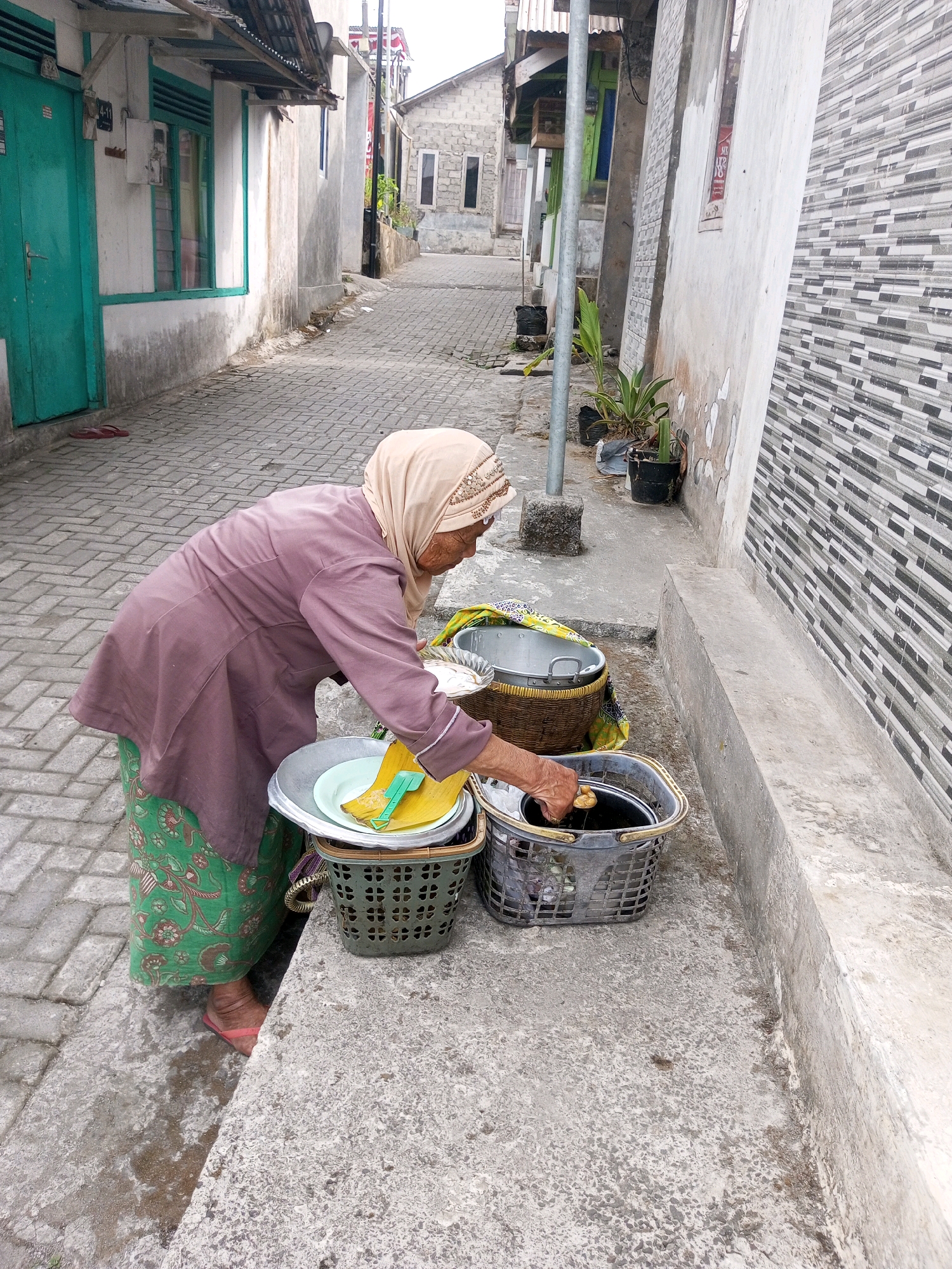 An elderly Indonesian woman wearing a light brown headscarf and a muted purple tunic over a green patterned skirt is seen from the side, facing away from the viewer, tending to a collection of baskets and basins on a paved pathway. The pathway is constructed of interlocking gray bricks. To her left, a building with a teal door and window frames is visible. The woman is actively engaged in an action, with her right hand reaching into a dark gray plastic basket containing a black bucket. Her left hand appears to be holding a yellow object over a large, silver basin. In front of her, there are two green plastic baskets, one containing a large silver basin with a light blue plate and a yellow object, and the other containing the black bucket. Behind these, a larger woven basket holds a silver basin. The environment appears to be an alleyway in Andongsili, Indonesia, with a narrow paved path flanked by buildings on either side. The building to the right features a textured wall of horizontally laid gray and black tiles. The overall scene depicts an everyday domestic activity taking place outdoors in a residential area.