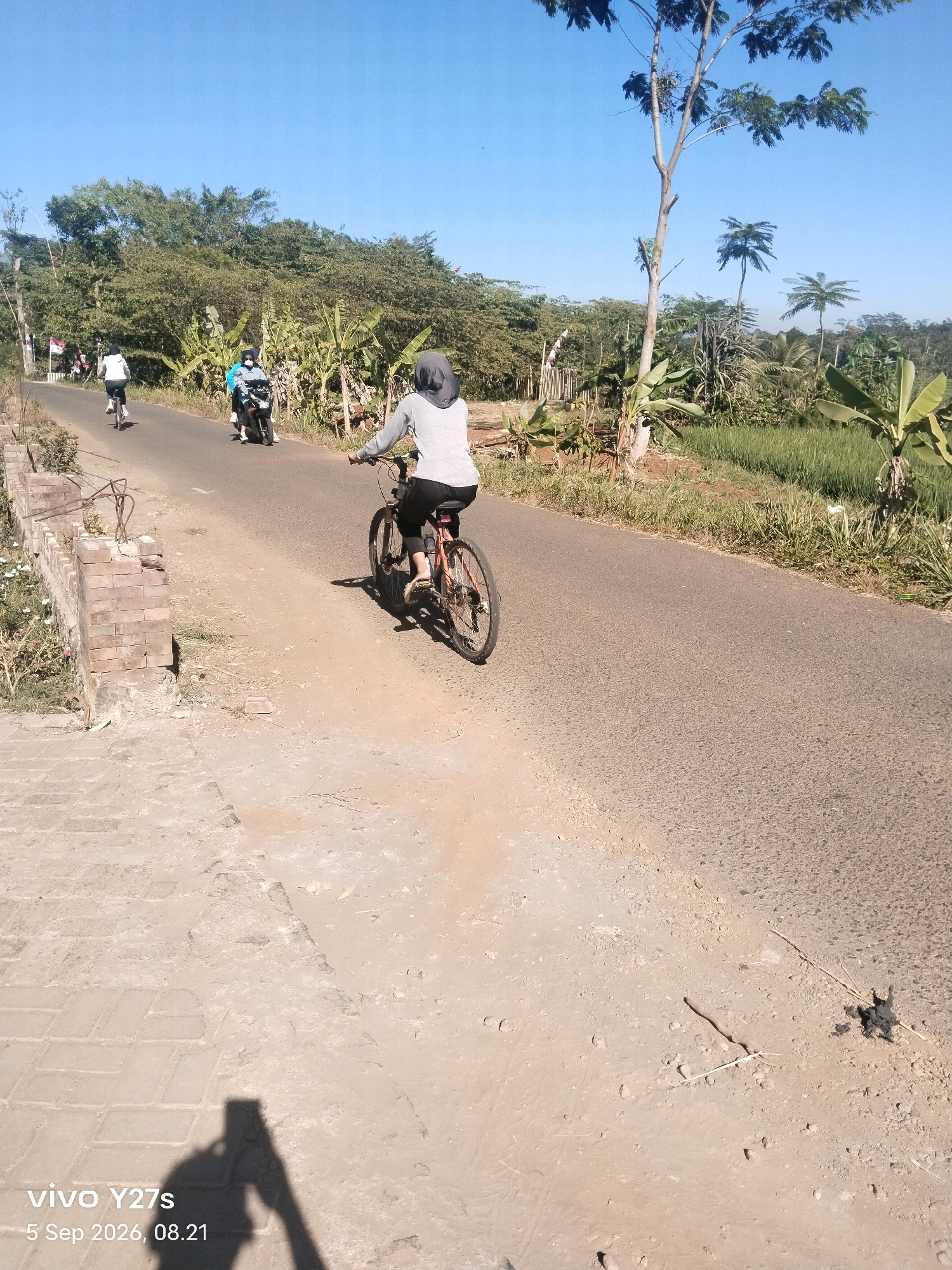 The image captures a clear, sunny day in a rural or semi-rural area, likely in Selojetis, Indonesia, given the presence of tropical vegetation like banana trees. The scene is dominated by a paved road, with a dirt path and a low brick structure on the left. In the foreground, a person is riding a bicycle from right to left, facing away from the camera. They are wearing a grey hooded top and black leggings. Further up the road, another person on a bicycle, wearing a white top and black leggings, is visible. Between the two cyclists, a person is riding a motorcycle, wearing a light blue top and a dark helmet. The background features lush green trees and foliage, suggesting a natural or agricultural setting. The sky is a vibrant blue, indicating good weather. The overall atmosphere is one of outdoor activity on a pleasant day.
