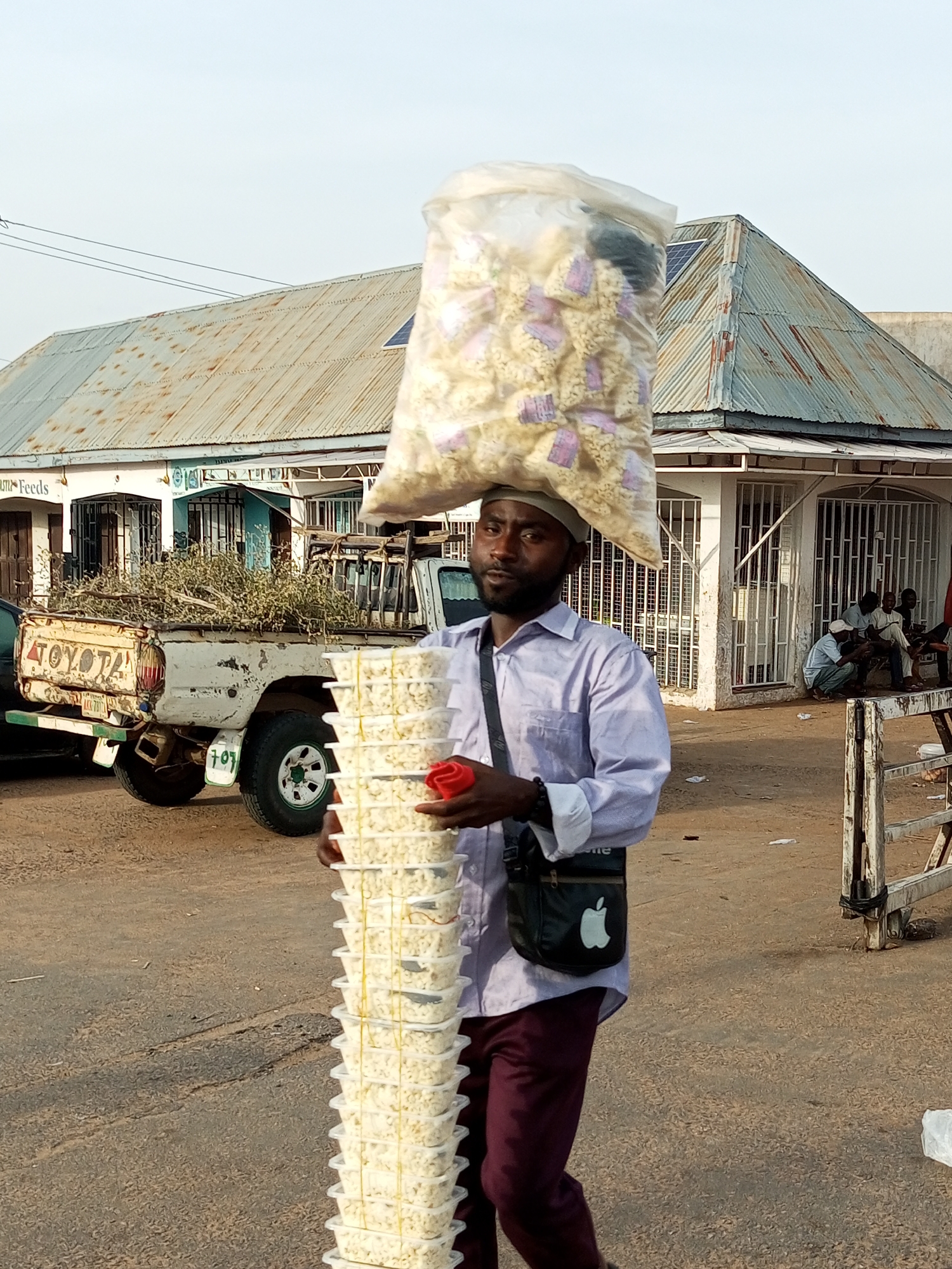 In the city of Gombe, Nigeria, a man is captured mid-stride, balancing a large plastic bag filled with what appears to be popcorn on his head. He is also carrying a tall stack of clear plastic containers, filled with the same food item, in his right arm. He is wearing a light purple long-sleeved shirt and burgundy trousers. A black crossbody bag with a white Apple logo is slung over his left shoulder. In the background, a white Toyota pickup truck is parked, its bed filled with dried vegetation. Several buildings with corrugated metal roofs are visible, some appearing to be shops with barred windows. A few people are seated or squatting in front of one of the buildings. The ground is a mix of dirt and asphalt, with some scattered litter. The sky is overcast.