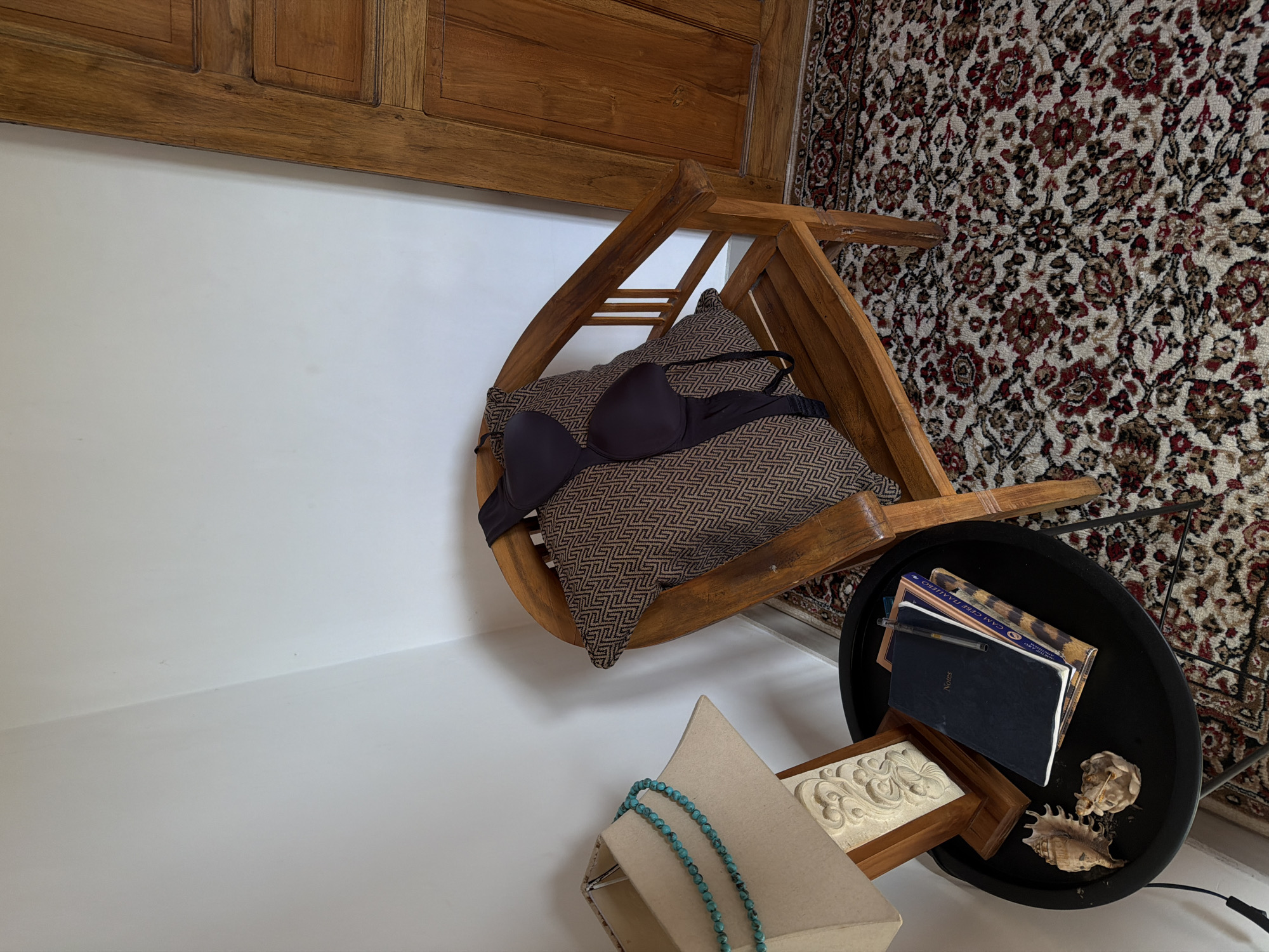 The image shows a bedroom or living area in North Kuta, Indonesia. There is a wooden headboard visible at the top of the frame, indicating the presence of a bed. A small wooden chair is overturned, with a patterned pillow on it and a black bra on top of the pillow. The chair is positioned close to a round black side table. On the side table, there are books, notebooks, and seashells. The floor is covered with a patterned rug. In the foreground, a jewelry display case with a turquoise necklace is visible. The room appears to be in disarray.