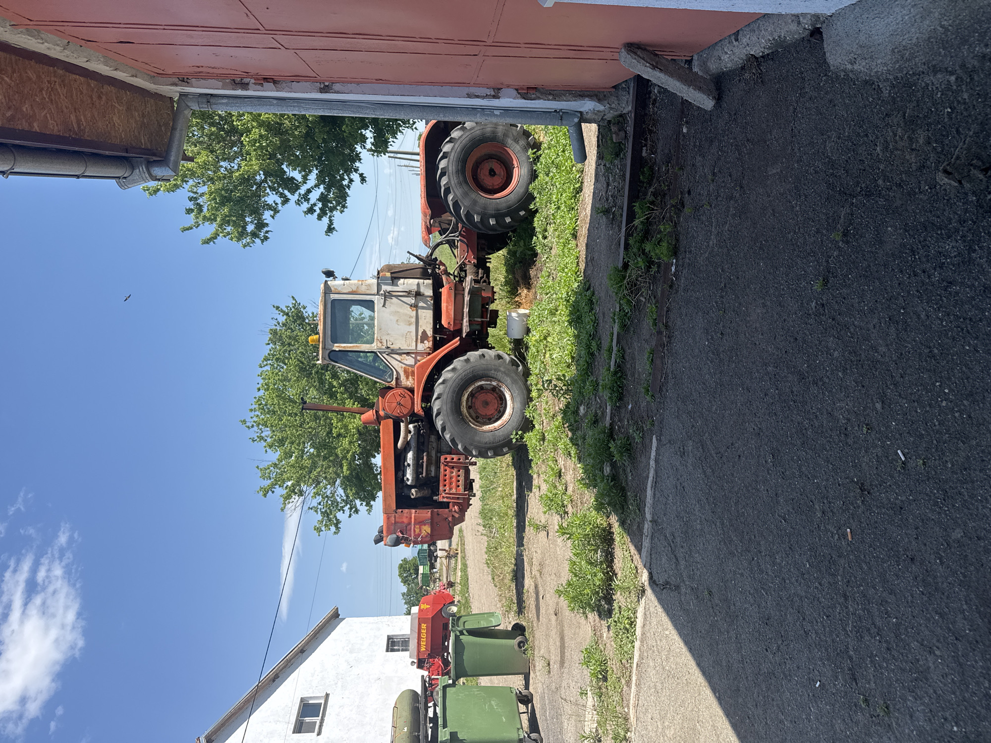 The image depicts a rural scene, likely on the outskirts of Sarnevo, Bulgaria, as the location was specified. An orange and white tractor is prominently featured, partially obscured by a doorway of a red building. The tractor's tires are large, and it appears to be well-worn. To the left of the tractor, outside the building, are green trash bins and a red agricultural machine, possibly a harvester. A white building is visible in the background on the left. The sky is clear blue with scattered trees in the scene. The ground is a mix of asphalt and grassy patches. The image appears to be taken on a sunny day.