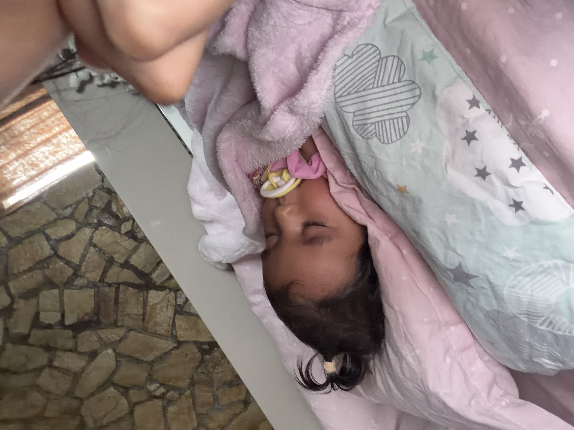 A baby is sleeping soundly in a crib with a pink blanket and a pacifier in its mouth. The crib is placed near a wall with a stone pattern and a window with sheer curtains. A person's hand is visible in the upper right corner of the image, suggesting someone is watching over the baby. The scene appears to be in a bedroom in Razhevo Konare, Bulgaria.