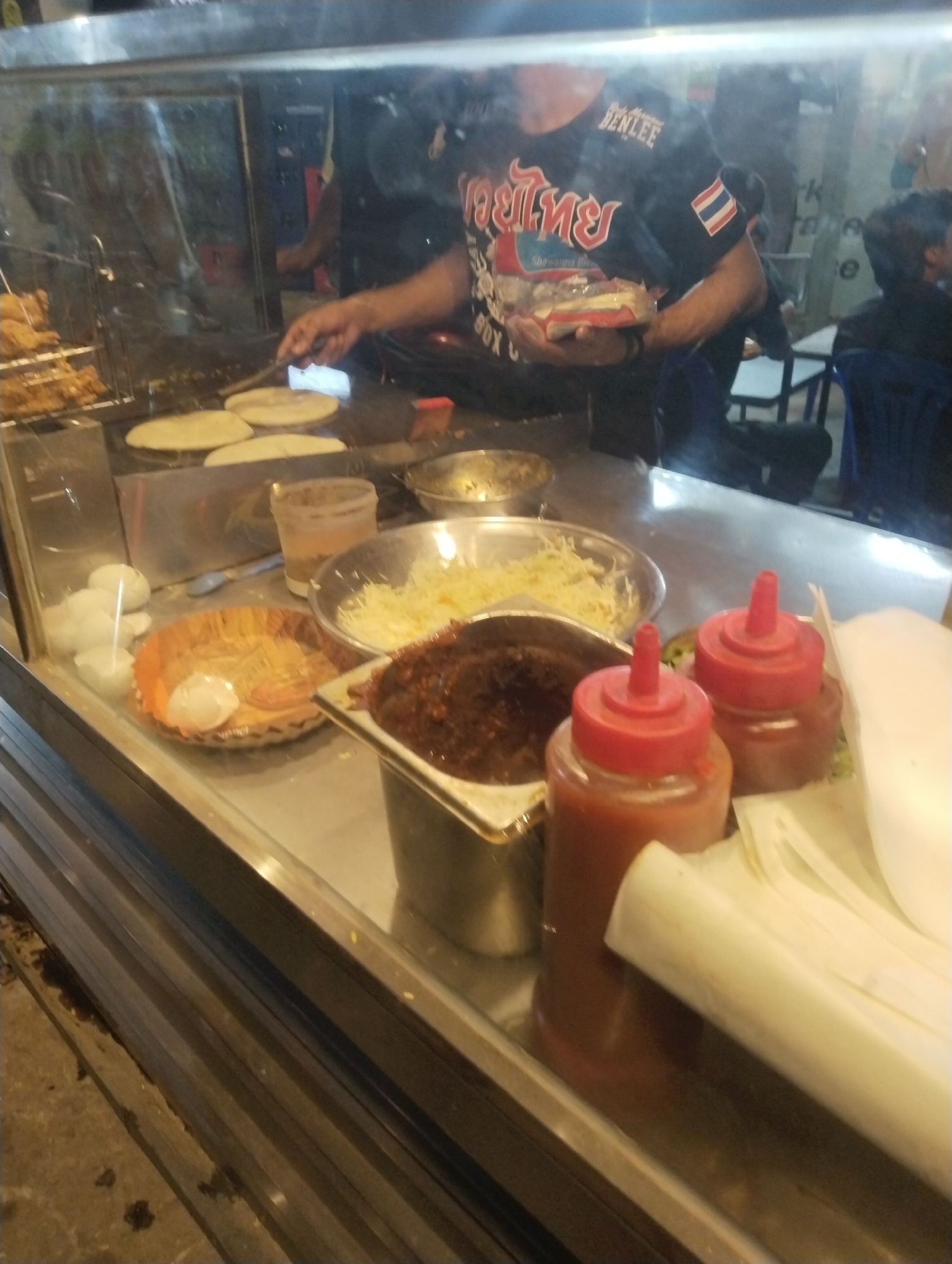 The image depicts a vibrant street food stall, likely operating in the evening given the artificial lighting and reflections. A male vendor is actively engaged in preparing food behind a glass partition. He wears a black t-shirt that prominently features Thai script ("มวยไทย," meaning Muay Thai), the word "BENLEE," and a Thai flag patch on the sleeve. In his left hand, he holds a white package, partially inscribed with "Shawarma Box," while his right hand uses a spatula to maneuver several flatbreads (resembling tortillas or parathas) cooking on a griddle.
On the counter in front of the vendor, various ingredients and condiments are neatly arranged. These include a large bowl of finely shredded white produce, possibly cabbage or cheese, and a metallic container holding a dark, rich sauce or relish. To the left, a rack displays several pieces of golden-brown fried chicken. Further left, an orange paper plate holds what appears to be boiled eggs. Two red-capped squeeze bottles, likely containing ketchup and chili sauce, are placed prominently on the counter, ready for use. The overall scene, set in Heir, Pakistan, suggests a popular food spot offering wraps or shawarmas, possibly with a fusion or international theme indicated by the vendor's attire and the "Shawarma Box" text.