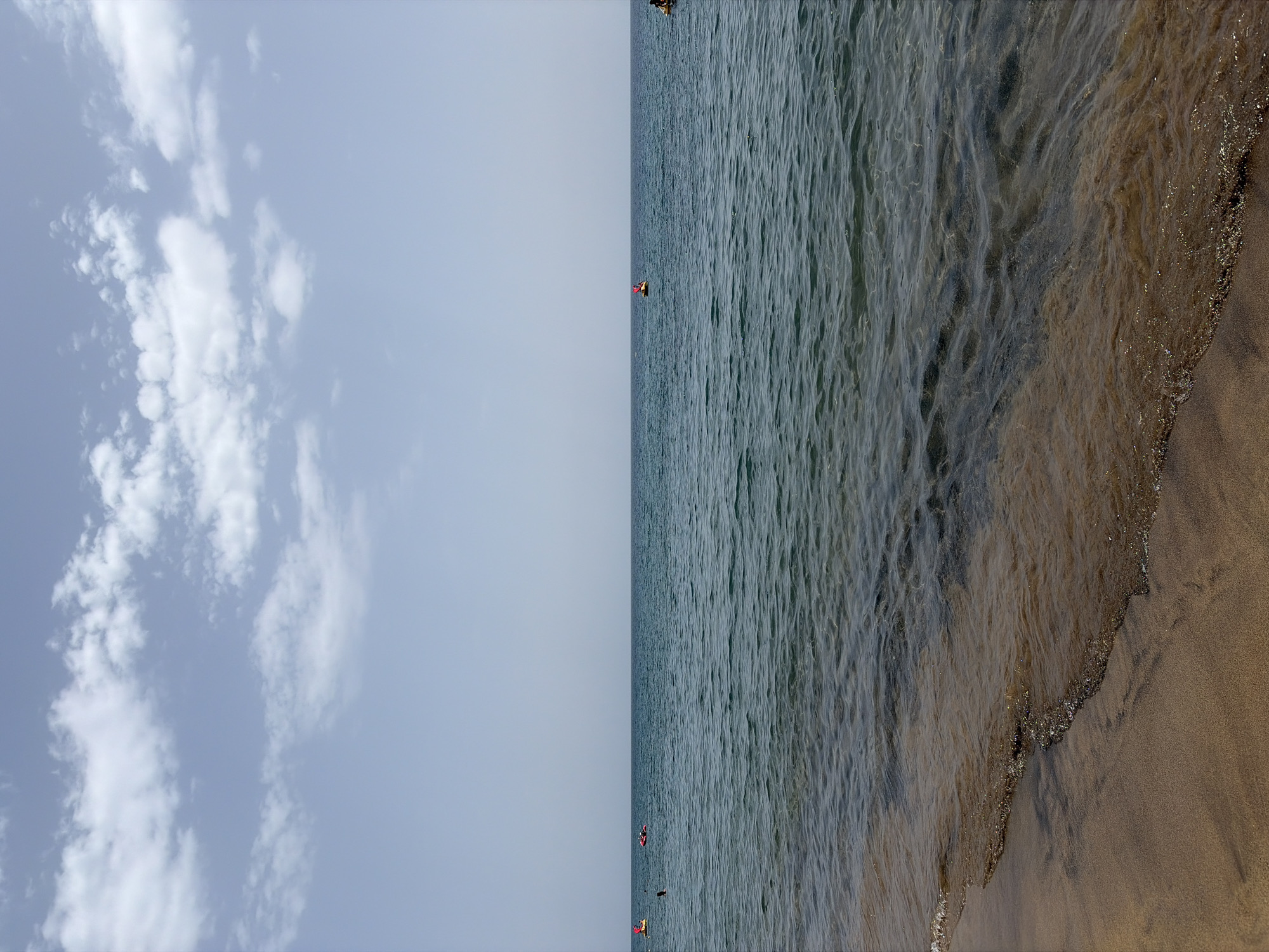 This image depicts a beach scene. The left side features a sky with scattered white clouds against a light blue backdrop. The right side showcases the ocean meeting the shoreline. Several small figures can be seen in the water, suggesting the presence of people engaging in water activities. The texture of the water is visible, with gentle ripples indicating slight movement. The sand on the beach appears to be dark in color. Given the context and mention of Puerto Del Carmen, Spain, it can be inferred that this scene is likely taken at Playa Grande, one of the main beaches in the city.