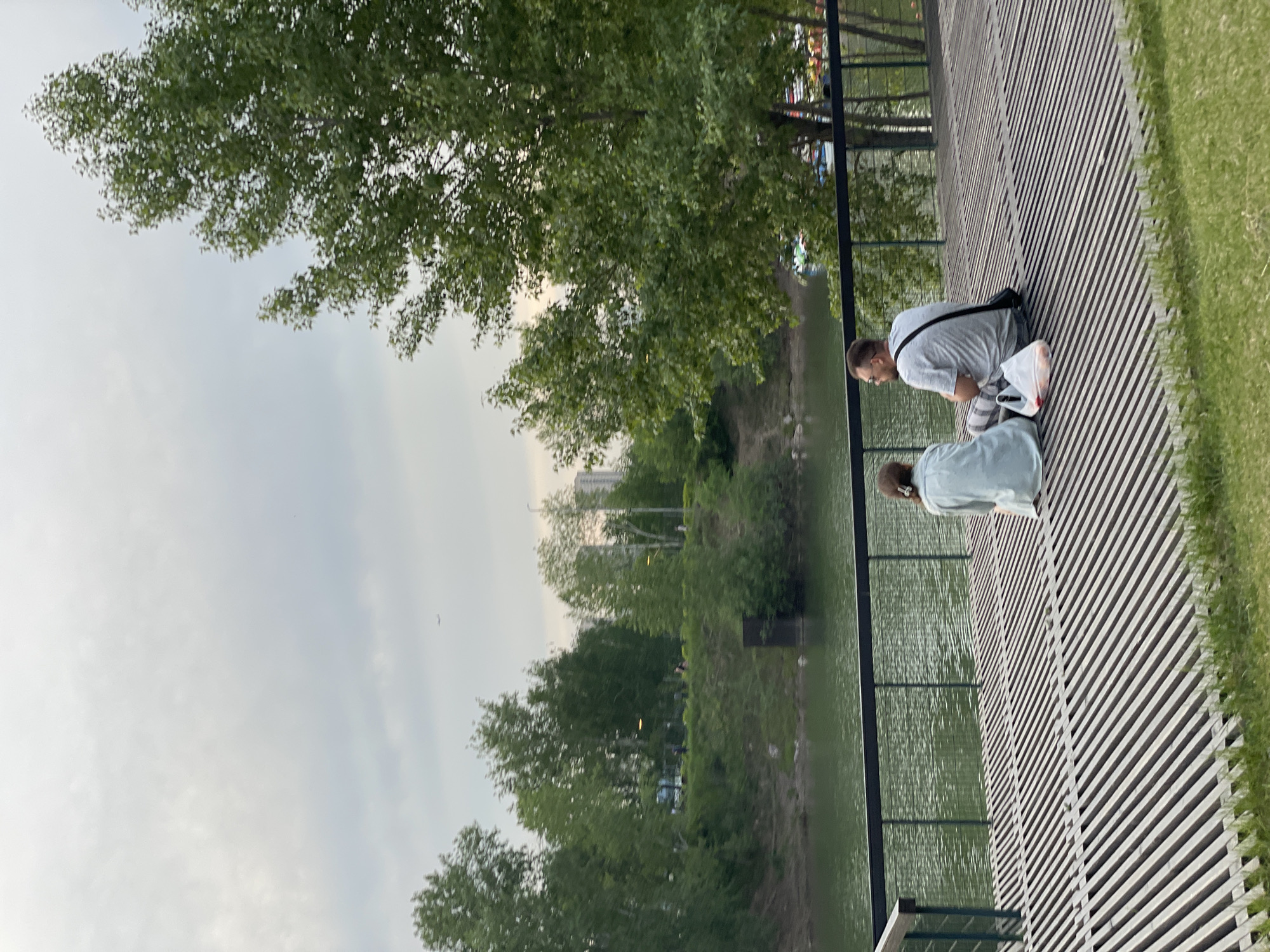 The image shows two people on a wooden boardwalk next to a body of water. The person in the foreground is wearing a light blue shirt and is facing away from the camera, looking towards the water. The person behind them is wearing a gray t-shirt and striped shorts, also looking towards the water. They are both in a crouched or kneeling position. A black bag is slung over the shoulder of the person in the gray shirt, and a white plastic bag is next to them on the boardwalk.
The setting appears to be a park or recreational area. Trees with lush green foliage line the bank of the water, and grass is visible on the right side of the boardwalk. The sky is overcast with some patches of lighter clouds, suggesting it might be a cloudy day or early morning/late evening. The water is calm and reflects the greenery and sky. The boardwalk is made of wooden planks laid diagonally. In the background, beyond the trees and water, there are hints of urban structures, including tall buildings. The overall mood is tranquil and observational. No visible text is present in the image. The location context is Krasnoyarsk, Russia.
