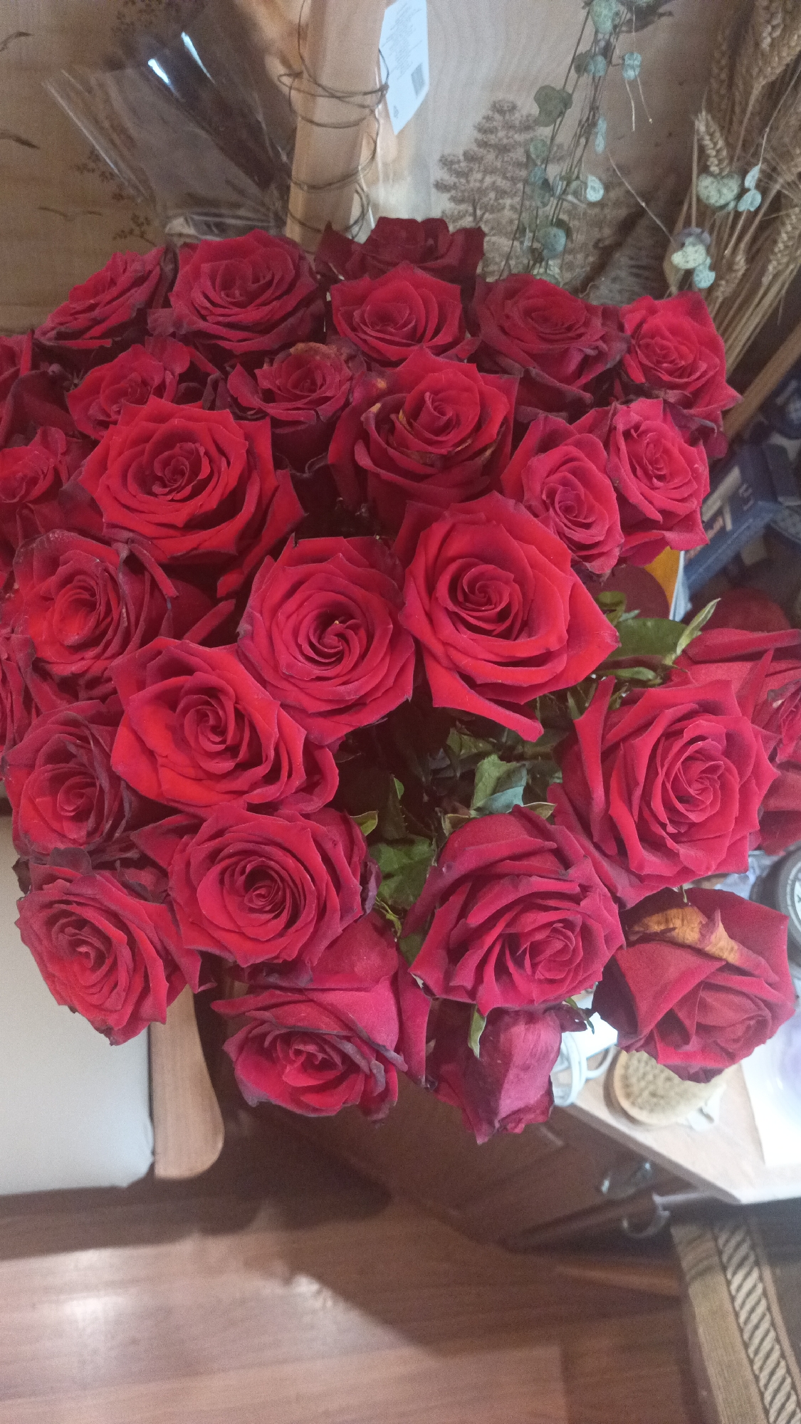 The image shows a bouquet of approximately 25 red roses in full bloom. The roses are densely packed together. Some green leaves are visible amidst the flowers. The bouquet is situated indoors, likely on a table or counter with a light brown wood-grain surface. In the background, there's a blurry arrangement that includes elements like wheat stalks and some decorative wrapping. Based on the query, the location of the scene is Donino, Russia.