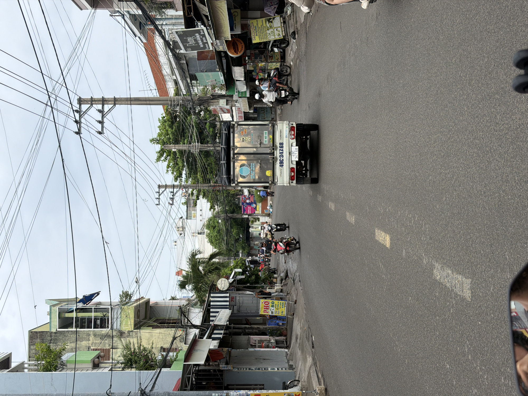 The image depicts a street scene in Nha Trang, Vietnam, likely in a commercial or residential area with a mix of buildings and overhead electrical wires. The road is paved and has white lane markings. On the left side of the street, there are multi-story buildings with awnings and signage, indicating shops or businesses. A yellow sign with Vietnamese text is visible. Several motorbikes are present, with some people riding them and others parked. A small white truck with a refrigerated cargo box is driving down the road, displaying the license plate "49C.347.68". Trees and vegetation are interspersed between the buildings. The sky is overcast. The perspective suggests the photo was taken from a moving vehicle, possibly a motorbike, as indicated by the presence of a side mirror in the lower left corner reflecting a person's arm and shoulder.