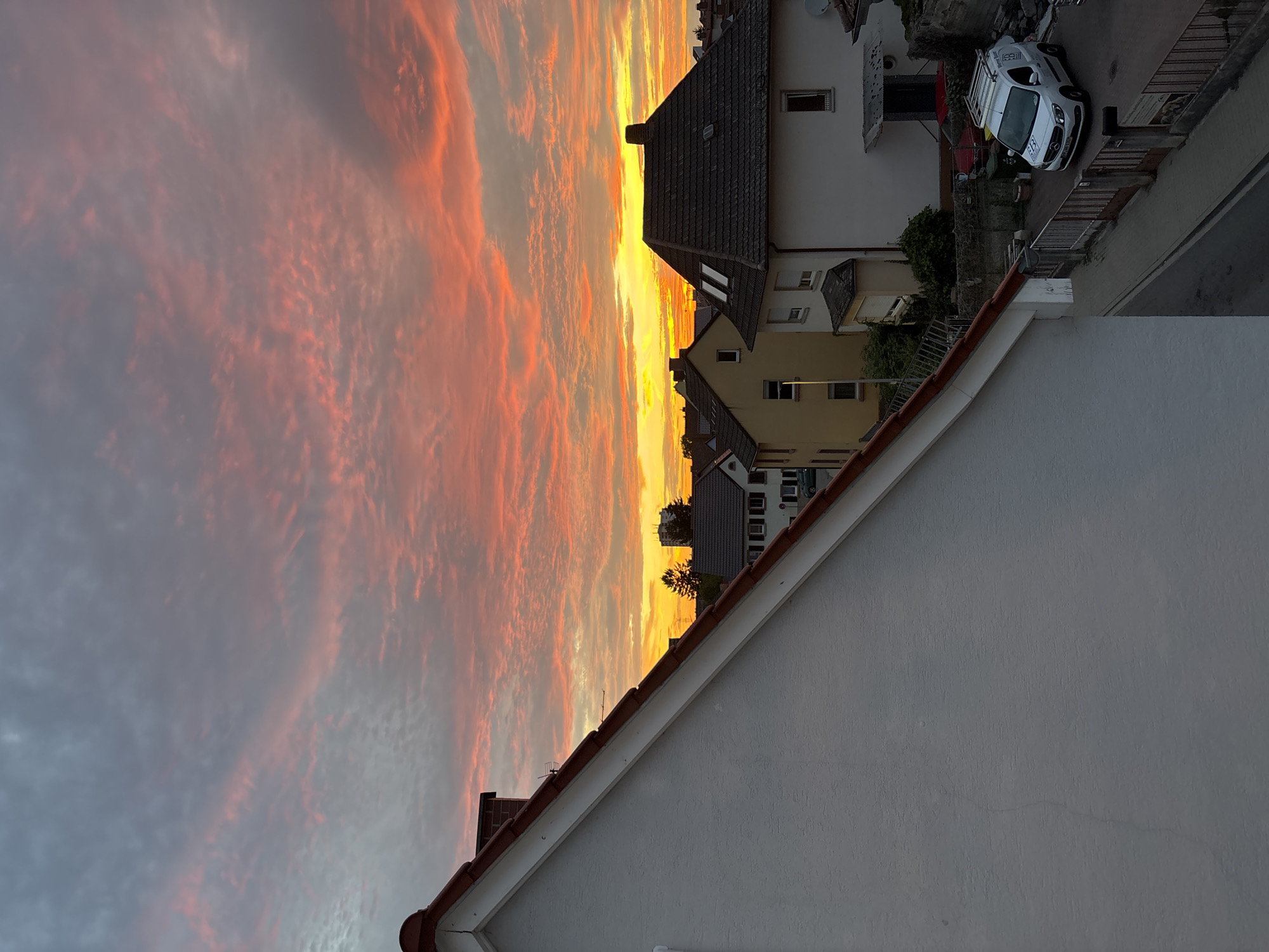 The image depicts a residential area in Viernheim, Germany, at sunset. The sky is filled with dramatic clouds painted in vibrant hues of pink, orange, and yellow, indicating the sun is low on the horizon. In the foreground, the angled roof and white textured wall of a house dominate the left side of the frame.
Beyond this house, a cluster of other residential buildings is visible. These include houses with dark tiled roofs and lighter colored facades, some with visible windows and chimneys. A prominent white van with "Bauer Solar" branding is parked in a driveway on the right side of the image. The van has a roof rack and appears to be a commercial vehicle. A paved road or driveway runs alongside the houses, with a metal fence bordering it. Greenery, including bushes and trees, is interspersed between the buildings, adding a touch of nature to the urban landscape. The overall scene suggests a peaceful evening in a suburban setting.