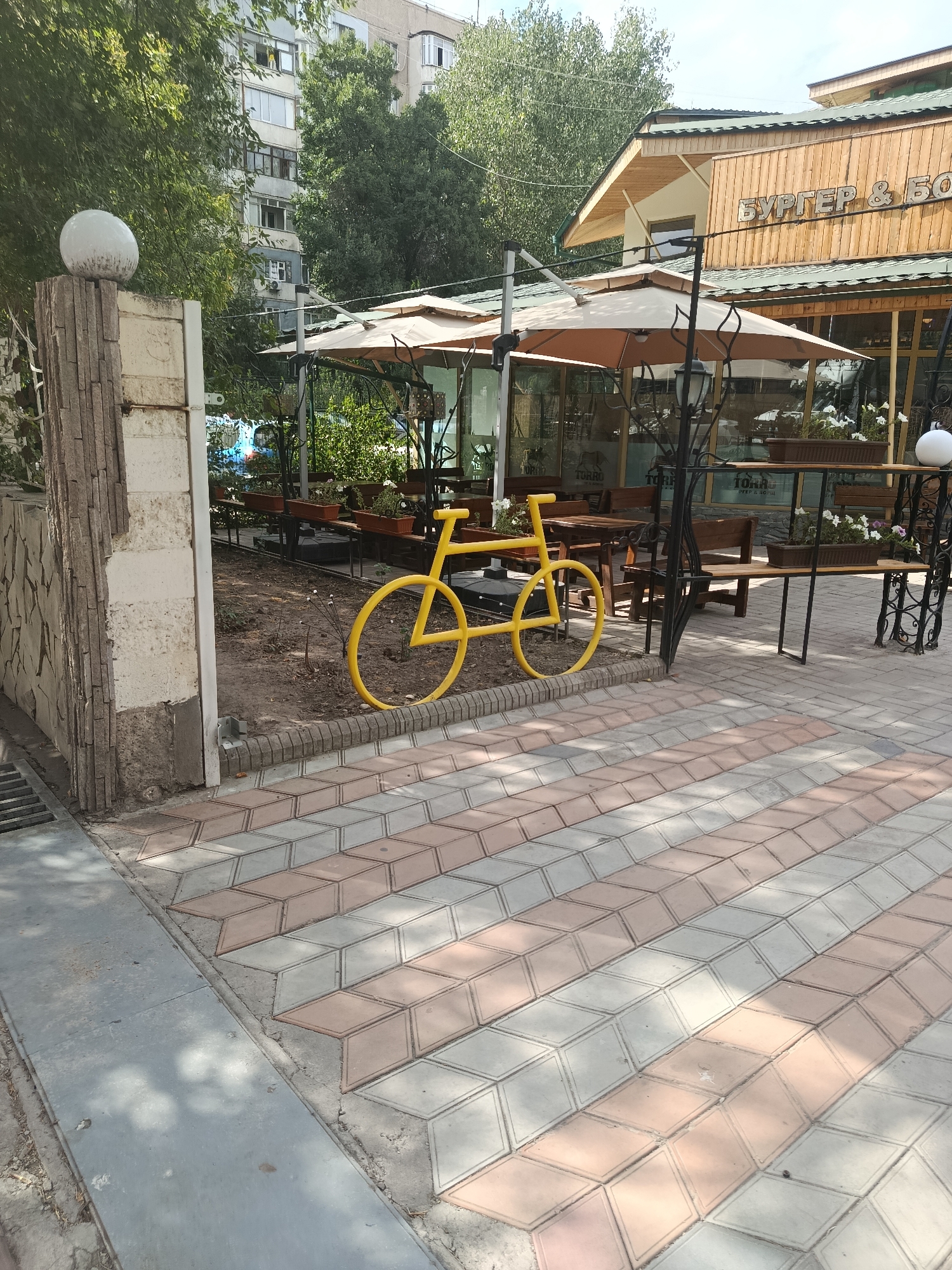 The image shows a yellow bicycle sculpture placed on a dirt area next to a sidewalk with diamond-shaped paving stones. Behind the bicycle is an outdoor seating area with tables and umbrellas, suggesting a cafe or restaurant. In the background, there is a multi-story building and trees. The building has signage that reads "БУРГЕР & БО" which is likely Cyrillic text for "Burger & Bo". The style of the building, the signage, and the general urban environment are consistent with Bishkek City, Kyrgyzstan. There are no people visible in the image. The overall scene depicts an outdoor space in an urban setting.