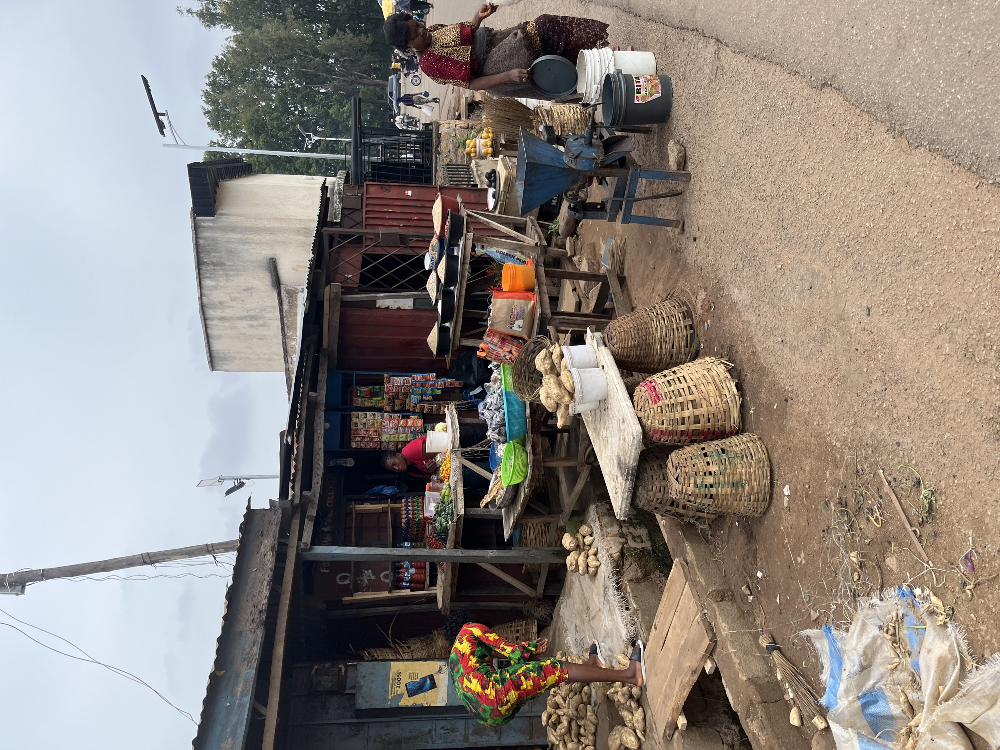 This image depicts a bustling market scene in Jos, Nigeria. In the foreground, a vendor in a vibrantly colored dress is bent over, examining root vegetables laid out on a rough mat. A pile of similar root vegetables is visible to her left, alongside a wooden plank. To the right, a woman in a patterned top and a dark wrap stands near a blue mechanical grinder, holding a dark lid over two buckets. Baskets of varying sizes are scattered around the market stalls, some filled with produce, others empty. The stalls themselves are constructed from corrugated metal and wood, displaying an assortment of goods including sacks of grain, bottles of condiments, eggs, and packaged items. In the background, other vendors and customers can be seen, along with motorcycles and cars on a paved road, suggesting a busy urban environment. The sky is overcast, indicating a potentially humid or rainy day.