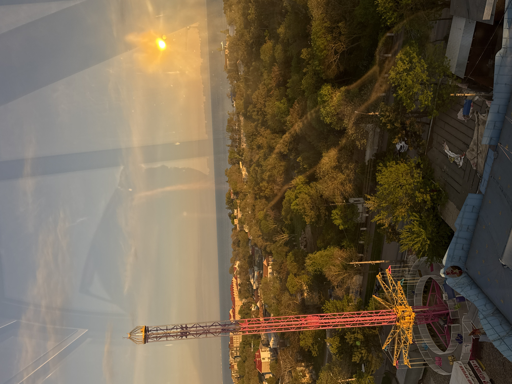The image depicts a view from a high vantage point, likely a Ferris wheel or similar structure, overlooking the city of Yevpatoriya, Ukraine. The Black Sea can be seen in the background, with the sun setting or rising over the water. A portion of a swing ride with a central tower painted in red and yellow is in the foreground. Below, rooftops of houses and buildings of the city can be seen, surrounded by trees. In the top right corner of the image, the roof of a building is visible. Some individuals are standing on the roof.