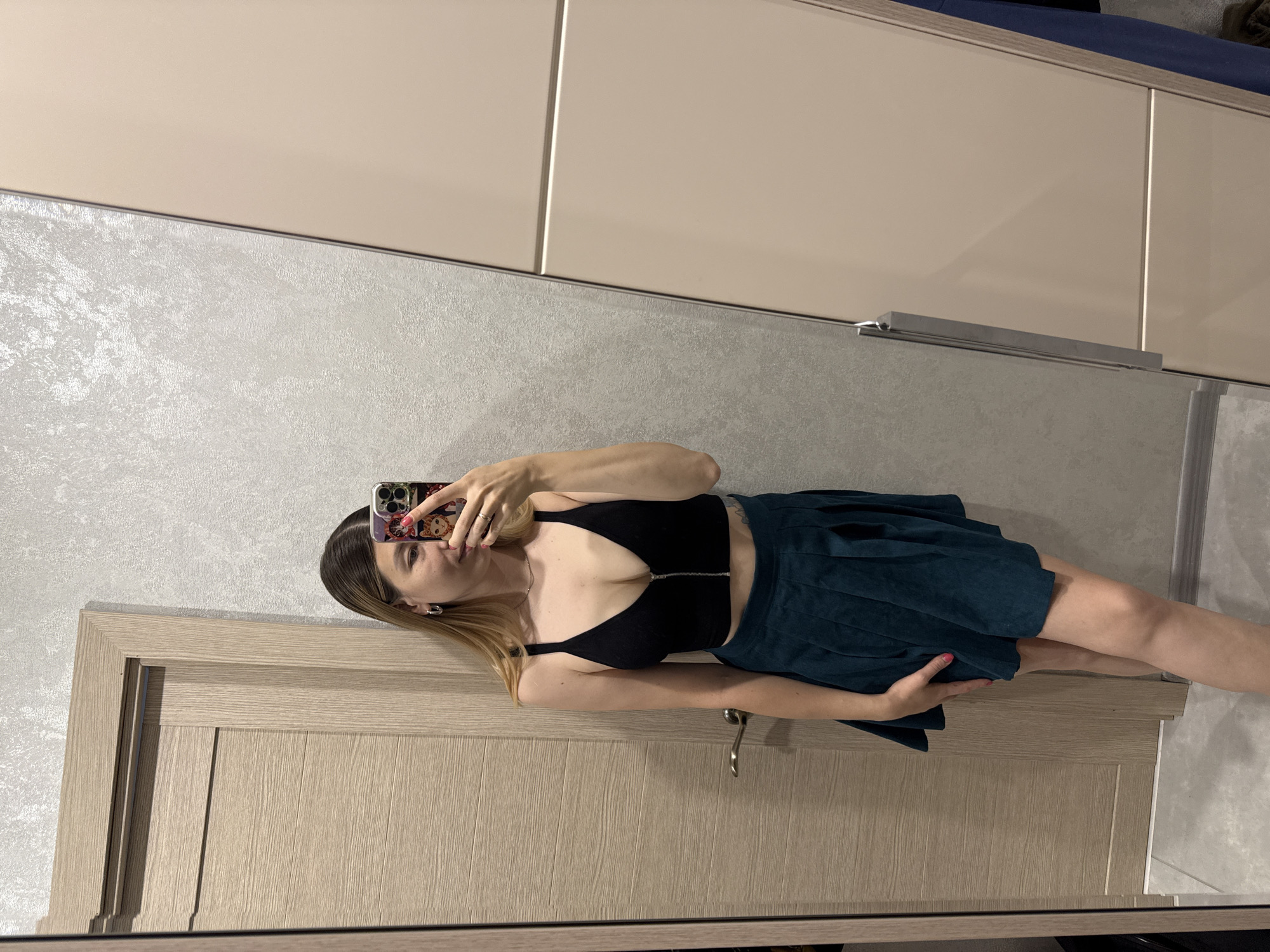 In Moscow, Russia, a young woman with blonde hair is taking a mirror selfie. She is wearing a black crop top with a zipper and a dark green pleated skirt. She is standing in front of a wooden door and a closet. The wall behind her is textured and light gray. Her phone, with a colorful case, is held up to capture the reflection.