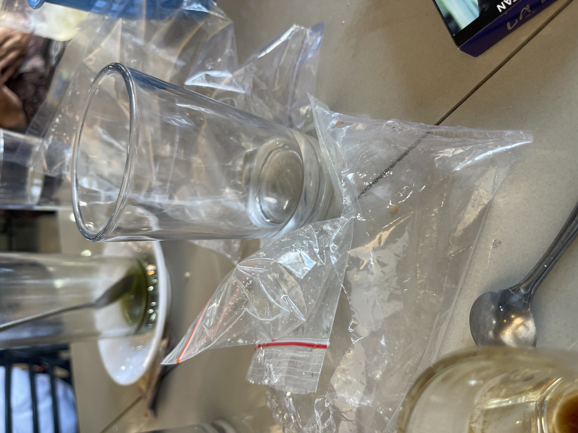 In the city of Bireuenmeunasahdayah, Indonesia, a table surface is cluttered with used drinking glasses, discarded plastic packaging, and a spoon. One glass, partially filled with a dark green liquid, sits on a white saucer. Next to it, an empty glass stands prominently. Several crumpled clear plastic bags are strewn across the table, some appearing to have been used for food or drink packaging. A metal spoon rests near the bottom left of the frame, partially obscured by a glass. In the background, a person's hands can be seen holding a patterned cloth, suggesting a dining or social setting. A blue object, possibly a bottle, is visible in the upper right corner. The overall scene depicts the aftermath of a meal or beverage consumption.