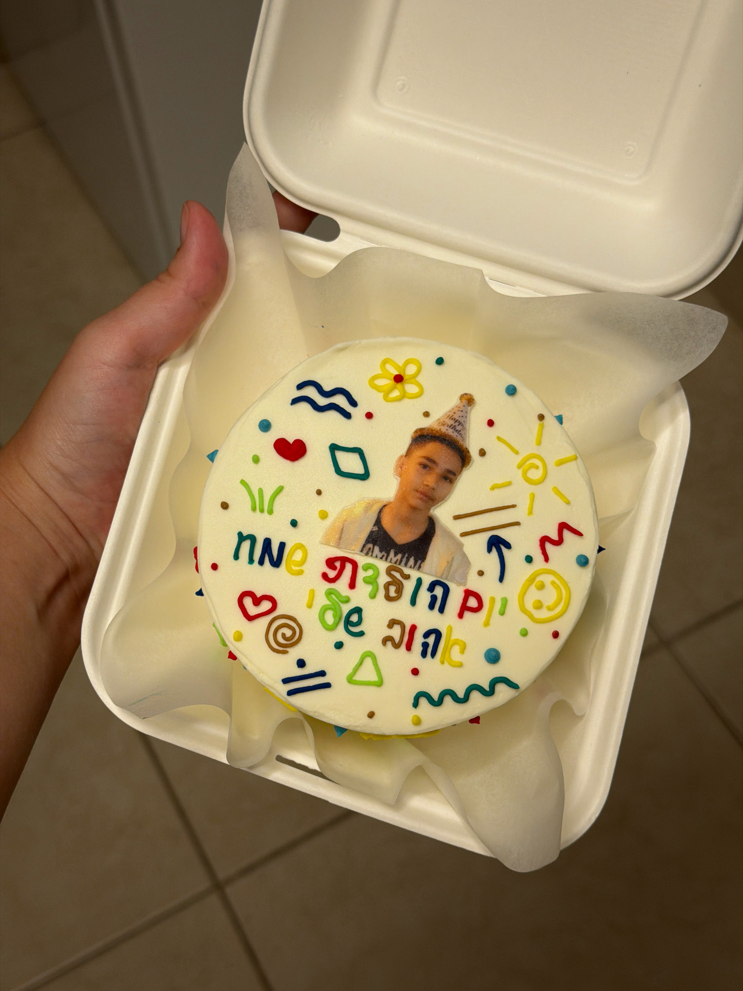 A person is holding a white takeout container with a white circular cake inside. The cake is decorated with colorful frosting in the shape of various symbols, including a flower, a heart, a sun, waves, and geometric shapes. A small edible photo of a young person wearing a birthday hat is placed on the cake. The background consists of a tiled floor and a partial view of a light-colored wall. The scene appears to be indoors, potentially a kitchen or dining area. There is no information to suggest this image is located in Tel-Aviv, Israel.