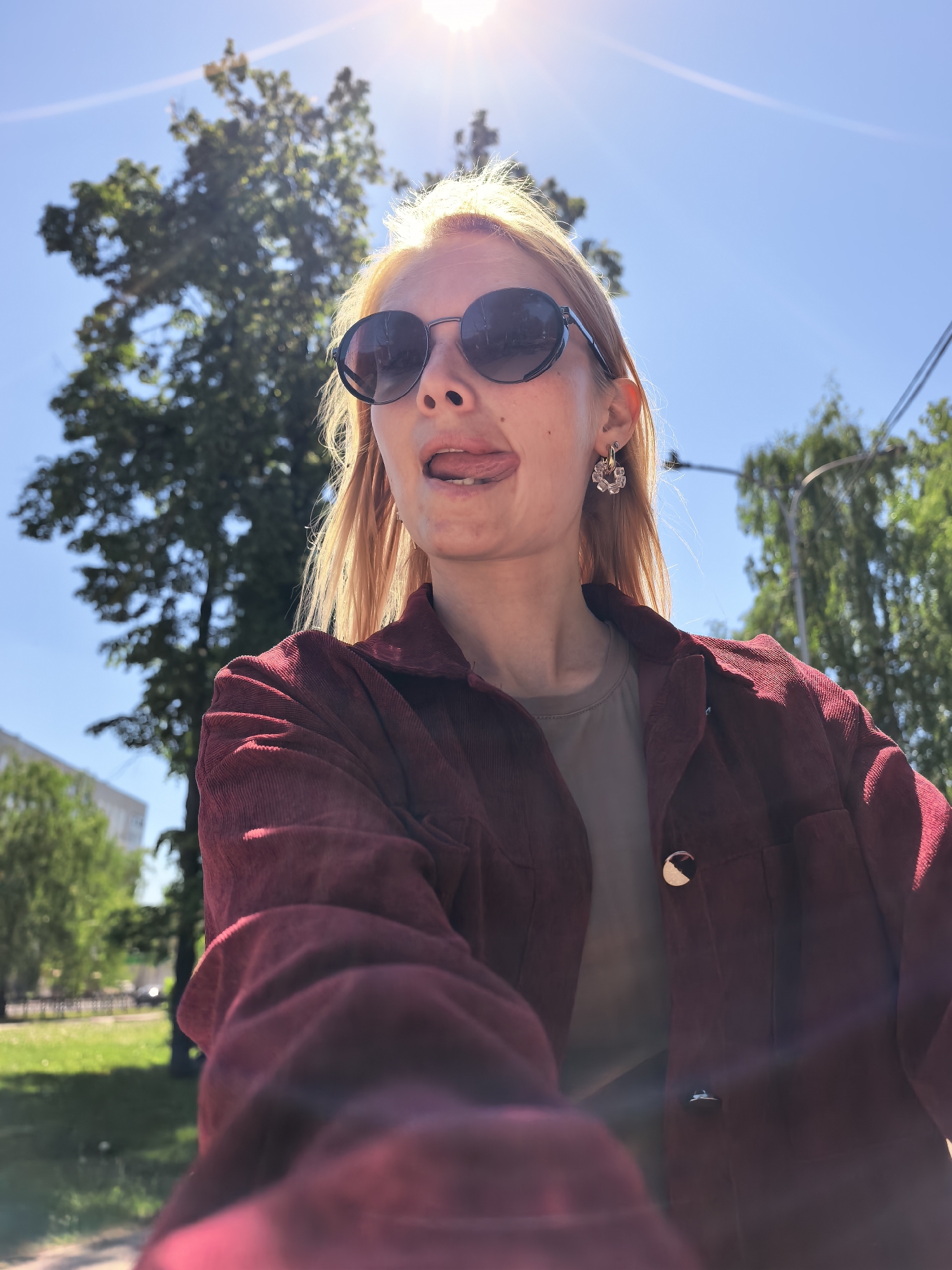 A woman with blonde hair and sunglasses sticks her tongue out in a selfie. She wears a red corduroy jacket over a grey t-shirt. Her earrings are visible. In the background, there is a clear blue sky with the sun shining brightly. There are green trees and a streetlight. The location is Sterlitamak, Russia.
