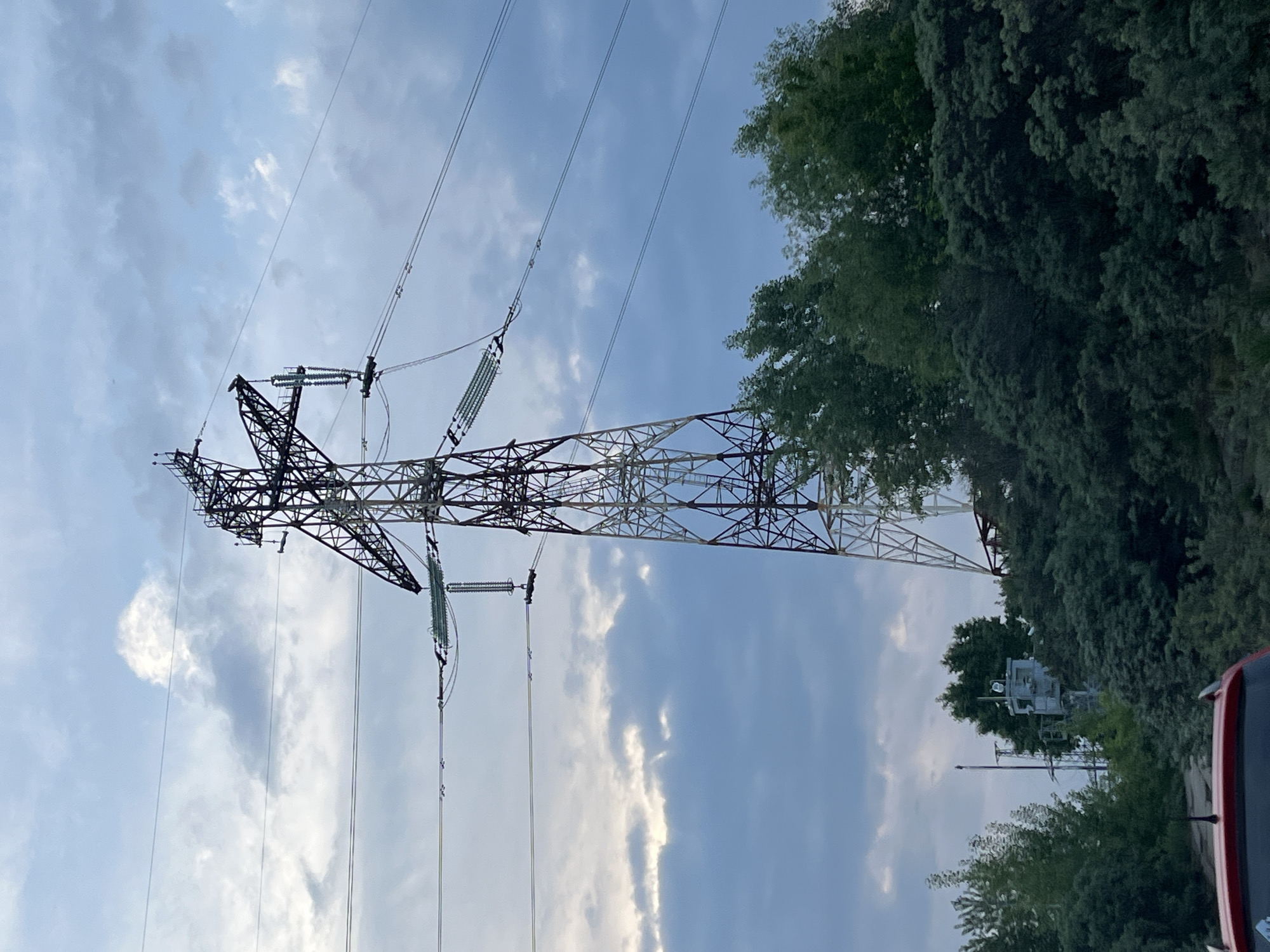 The image captures a towering electrical transmission pylon set against a partly cloudy sky. The dominant subject is a tall, intricate lattice-work steel structure, typical of a high-voltage power line tower. The tower features multiple cross-arms from which numerous power lines extend horizontally, supported by long strings of light green insulators.
The sky is a mix of light blue and white, with scattered, soft cumulus clouds visible in the background. Along the right side of the frame, a dense line of lush green trees partially obscures the lower section of the tower and the horizon.
In the bottom right corner, the top portion of a red vehicle, likely a car or SUV, is visible, including its antenna. Behind the trees, a grey structure resembling a utility box or communication equipment mounted on a pole can be discerned.
The scene depicts a static outdoor environment. It appears to be daytime, likely late afternoon, given the ambient light and the nature of the clouds, with no harsh shadows. No specific activity or people are present. There is no visible text in the image. The location is contextualized as Chaikovsky, Russia.
