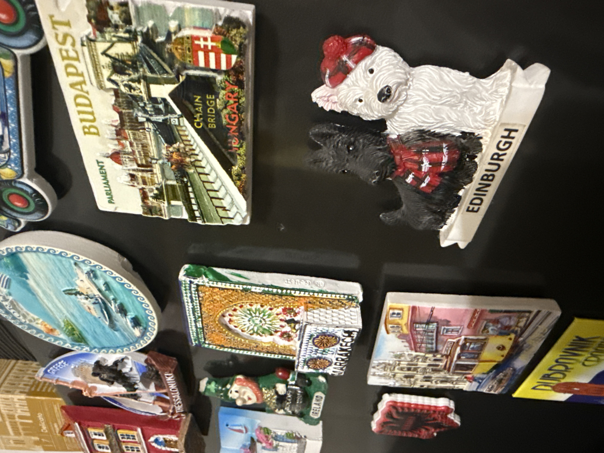The image shows a collection of souvenir magnets attached to a black surface, likely a refrigerator. The magnets depict various locations, including:
* "BUDAPEST," featuring the Hungarian Parliament and Chain Bridge.
* A pair of dogs with "EDINBURGH" written on the base.
* "GREECE" and "THESSALONIKI."
* "MARRAKECH."
* "IRELAND."
* "DUBROVNIK."
The magnets are oriented at different angles. There is no indication of the city of Cheshnegirovo, Bulgaria in the image.