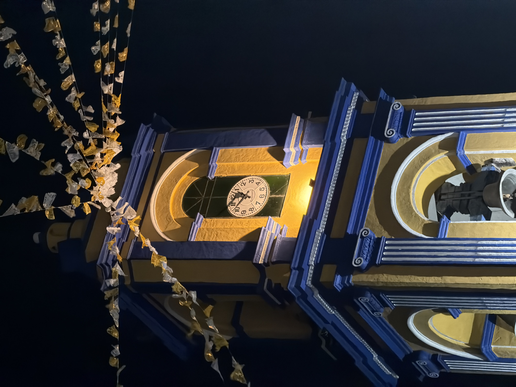 The image shows a portion of the clock tower of the San Andrés Cholula municipal palace, located in the city of San Andrés Cholula, Mexico. The structure is painted in a yellow and blue color scheme, and the clock face is visible and appears to be lit from behind. Decorative streamers hang in the foreground. The image was taken at night, as the background is dark.