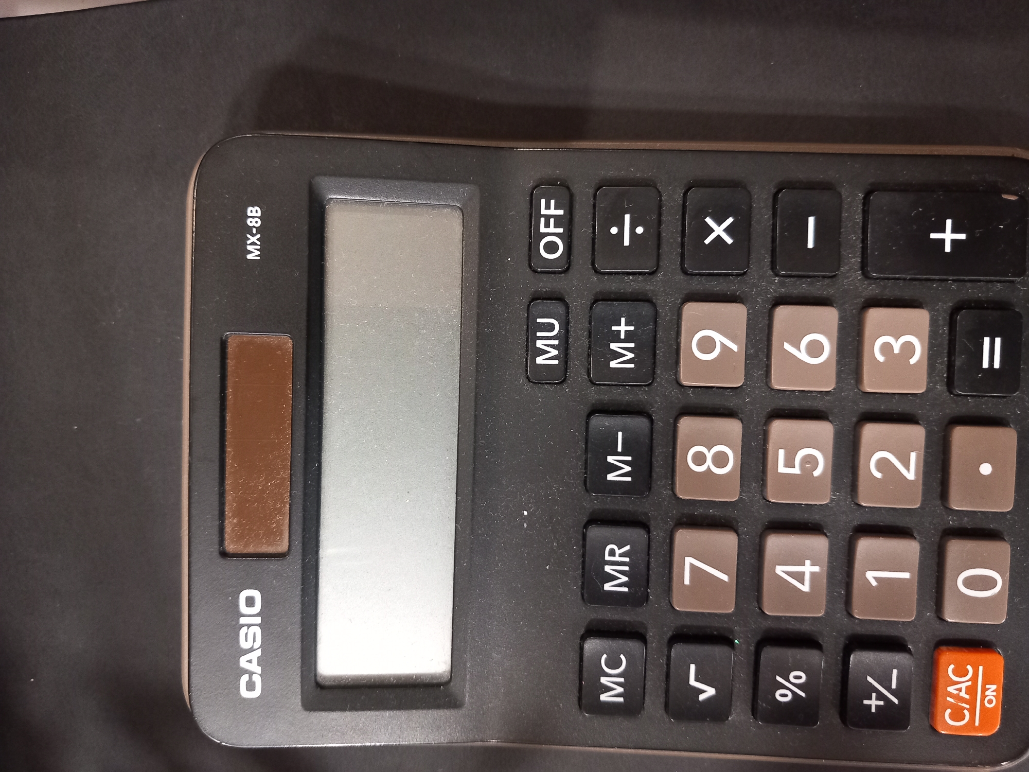 The image shows a close-up view of a black CASIO MX-8B calculator resting on a dark, textured surface. The calculator has a large, gray display screen at the top. Below the screen are rows of buttons. The top row of buttons includes "MU" and "OFF". The next row includes "M+", a division symbol, and a multiplication symbol. The following rows contain numbered buttons from 0 to 9, along with other function buttons such as a square root symbol, a percentage symbol, a plus/minus symbol, a decimal point, a minus symbol, and a plus symbol. The bottom left corner features an orange "C/AC ON" button. The calculator appears to be in good condition, with no visible damage. The lighting suggests an indoor environment, possibly a desk or table. The scene is static and no people or actions are depicted.
