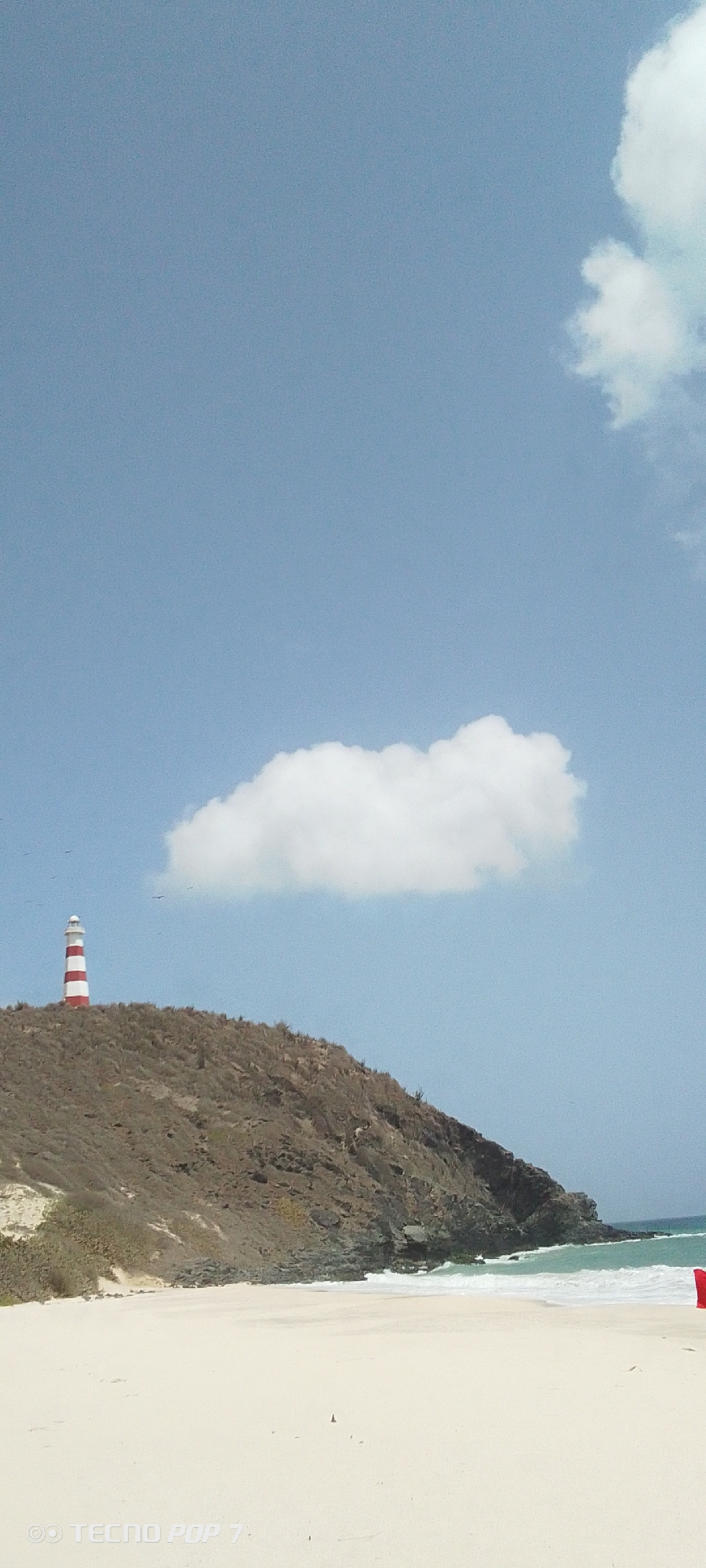 A vertical outdoor shot reveals the Macanao Lighthouse, situated on the arid promontory in Pedro González, Venezuela. The lighthouse is a red and white striped structure. A large white cloud sits in the sky above it. The foreground depicts a sandy beach and the ocean's edge. The sky is a light blue hue with a few other scattered clouds.