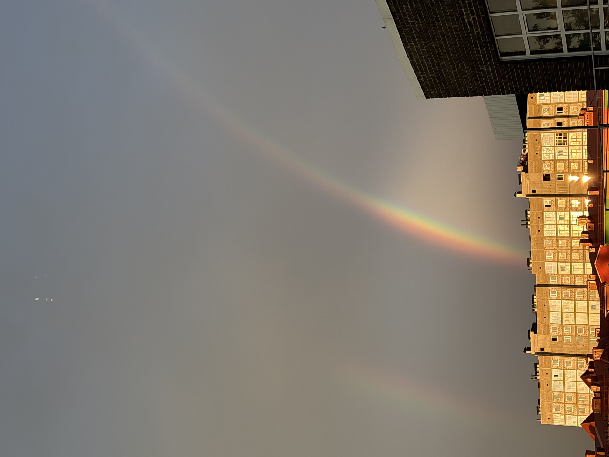 The image shows a sky with a prominent double rainbow arching over a cityscape. In the foreground, there are rooftops of buildings. Behind them, a large, light-colored apartment building with many windows is visible, illuminated by the warm light of the setting sun. To the right, a dark brick building with large windows stands out. The sky is overcast, with the sun's rays breaking through, creating the rainbow effect. The scene appears to be set in the city of Mikhaylovsk, Russia, given the context. There are no people or discernible actions depicted. The environment is urban.