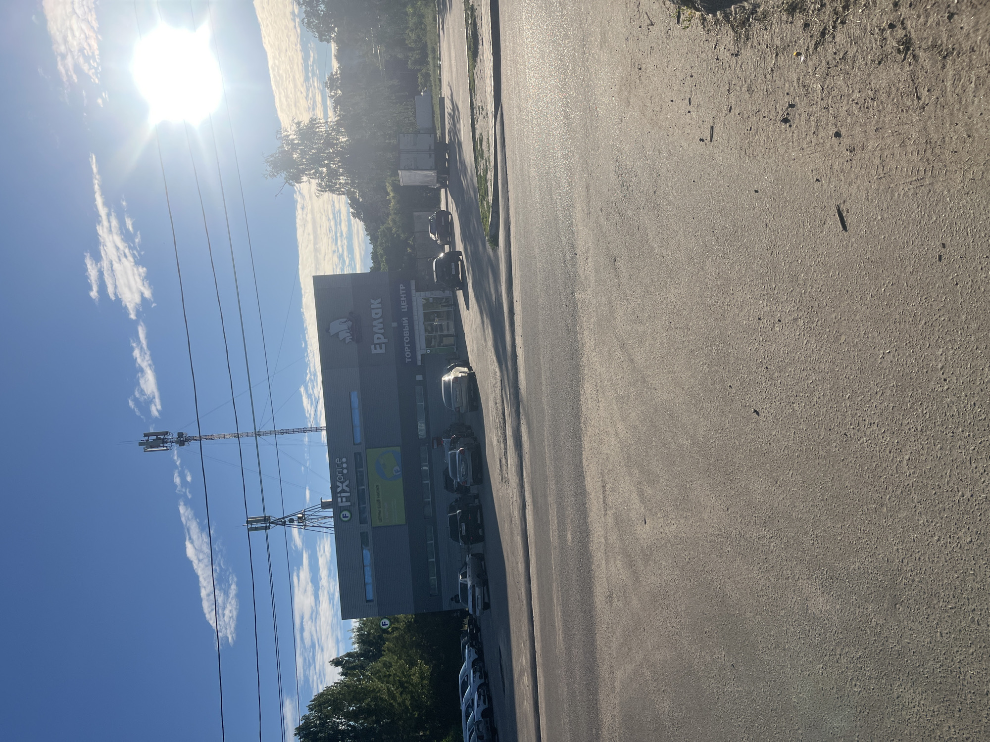 The image shows a parking lot in front of a commercial building. The building has two signs, one reading "FIX price" and another with the Cyrillic text "Ермак" and "ТОРГОВЫЙ ЦЕНТР" (Shopping Center). There are several cars parked in the lot, and a truck is visible in the background. The sky is blue with some clouds and the sun is shining brightly. The scene appears to be in a suburban or commercial area. This location is in Voronezh, Russia, specifically on Pervomayskaya Street, in front of the "Ermak" shopping center, which houses a "Fix Price" store.
