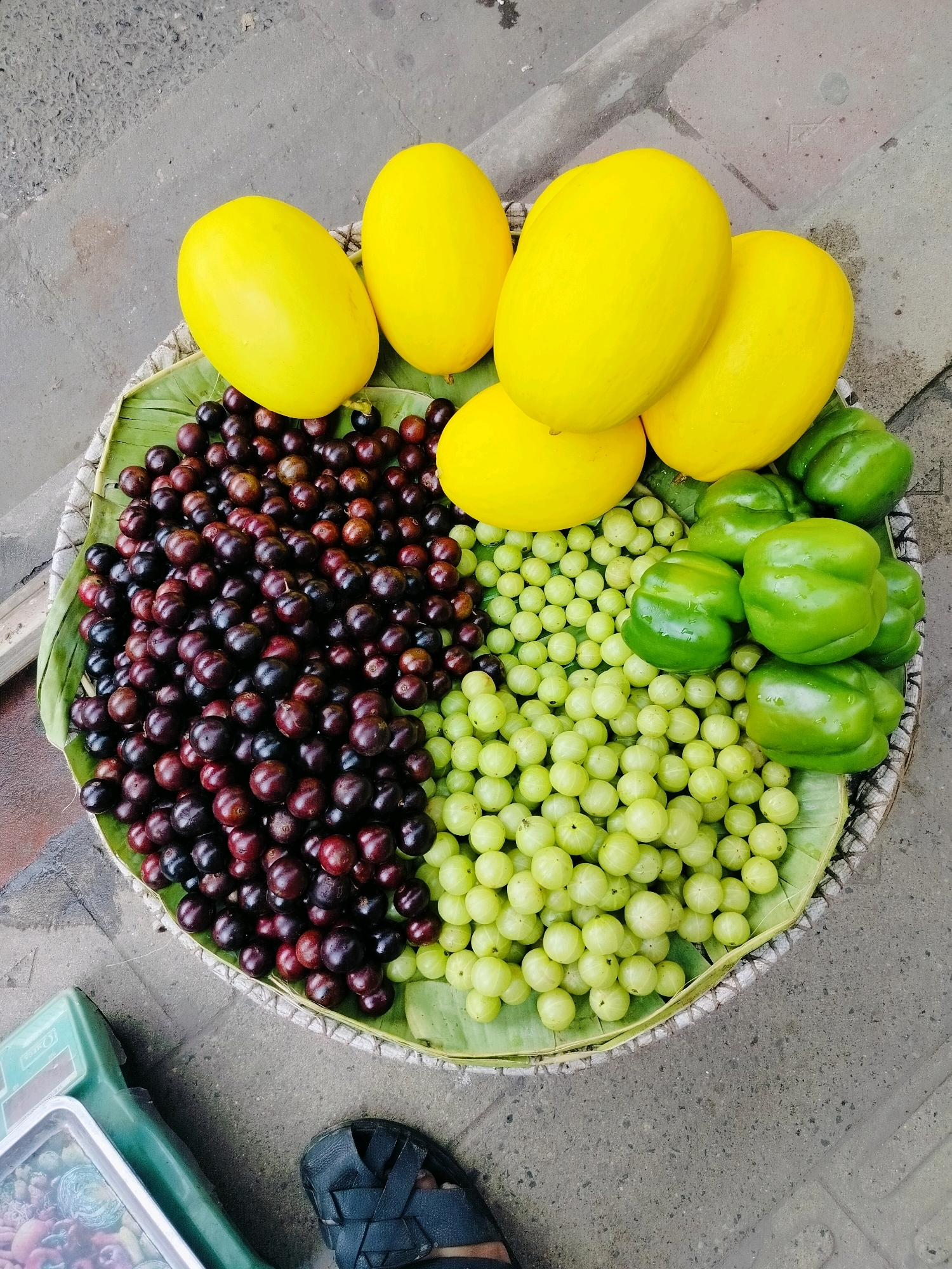 The image depicts a woven basket filled with various fruits and vegetables, placed on a gray, paved surface which is likely a street or sidewalk. The basket is lined with large green leaves, likely banana leaves.
Dominating the basket are several bright yellow, oval-shaped fruits, possibly melons or a similar variety. To one side of the basket are dark, small, round fruits, likely grapes or a type of berry, with a deep reddish-purple hue. Adjacent to these, filling another section of the basket, are small, light green, spherical fruits, which appear to be gooseberries. Finally, several vibrant green bell peppers are positioned in the upper right portion of the basket.
In the lower left corner of the image, a green and silver weighing scale is partially visible, with a transparent container on it holding more fruits, some of which are red and round. A black sandal with a textured sole is visible on the right side of the image, suggesting a person's presence.
The overall scene is consistent with a street vendor's stall in Dhaka, Bangladesh, where fresh produce is commonly displayed for sale on sidewalks.