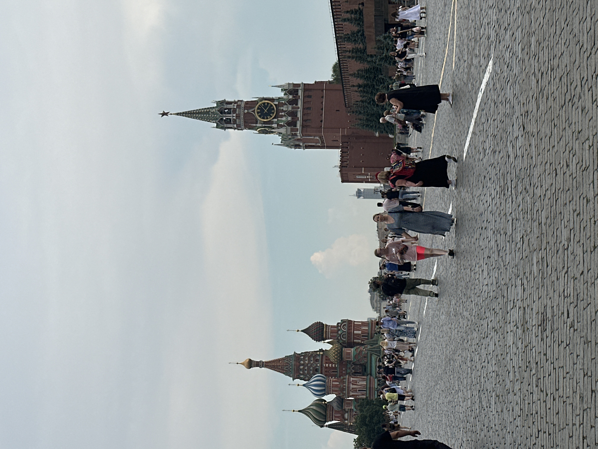 The image depicts a wide cobblestone plaza in front of two prominent landmarks. On the left is a vibrantly colored cathedral with multiple onion domes, featuring red and green hues, and topped with golden spires. To the right stands a large, imposing red brick clock tower, adorned with a green spire and a star at its apex. The sky above is overcast with scattered clouds. Numerous people are dispersed across the plaza, walking and standing. Several individuals are captured mid-stride, some in small groups, others walking alone. The architecture suggests a historical or cultural site in a European city, likely Russia given the architectural style. The ground is paved with an intricate pattern of grey cobblestones.