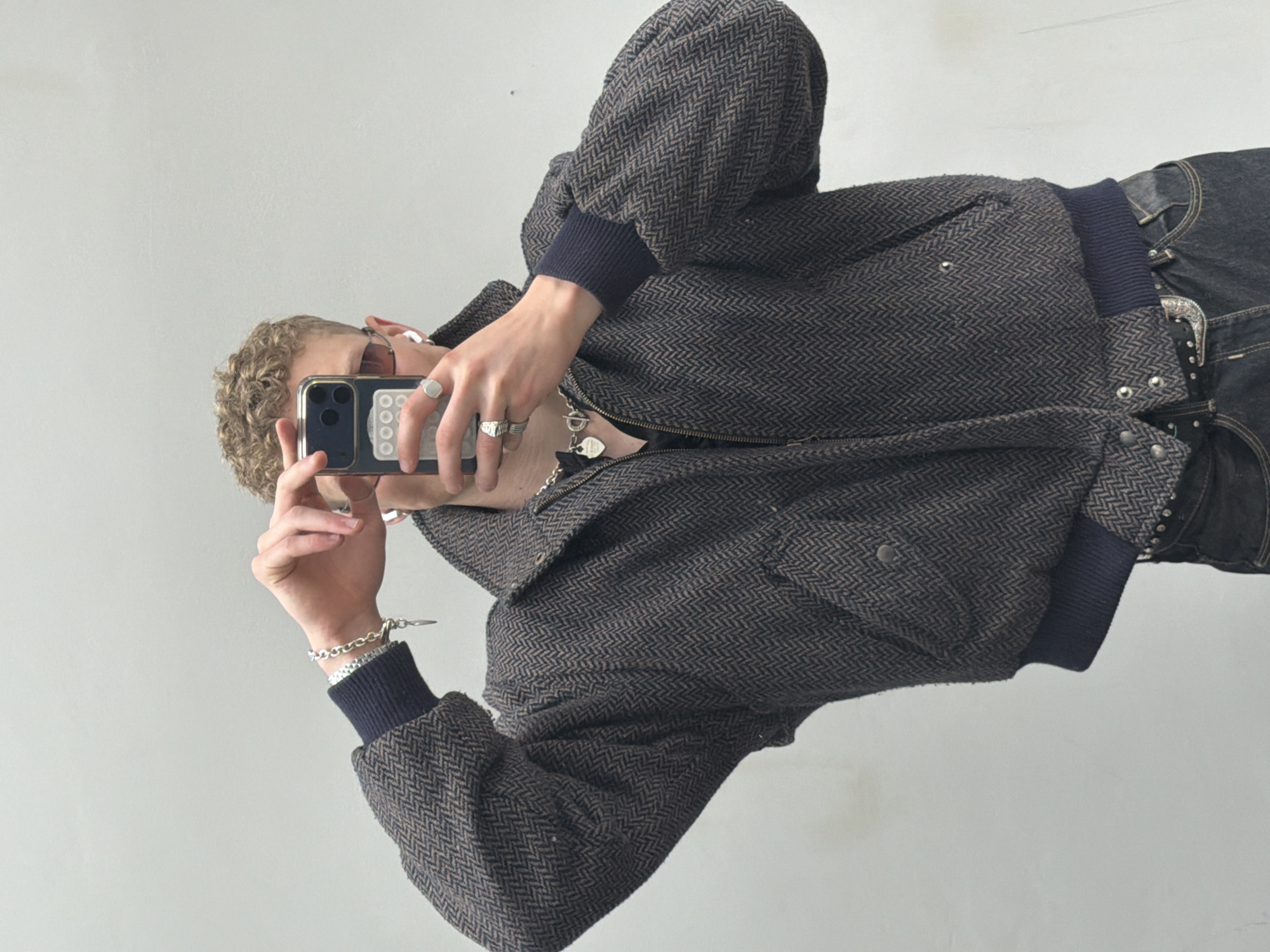 A person, likely male, with curly blonde hair, is depicted taking a mirror selfie or photograph with a smartphone, captured in a sideways orientation against a plain, light grey wall. The subject is wearing a dark grey or brown bomber-style jacket featuring a prominent herringbone pattern and dark blue ribbed cuffs. Dark jeans or trousers are visible, along with a belt adorned with a decorative silver buckle and studs.
The person's hands are decorated with multiple silver rings, including a large signet ring, and a silver chain bracelet is worn on the left wrist. A silver chain necklace with a small pendant is visible around the neck. Small white earbuds are placed in the ears, and sunglasses rest partially on the head, obscured by the phone. The smartphone, possibly an iPhone, is held with both hands and housed in a dark case that appears to have a card holder attachment with small white circular or square details on its back. The face is largely hidden by the phone, making emotional expression undeterminable. The setting is indoors, with a neutral background, offering no specific location cues.
