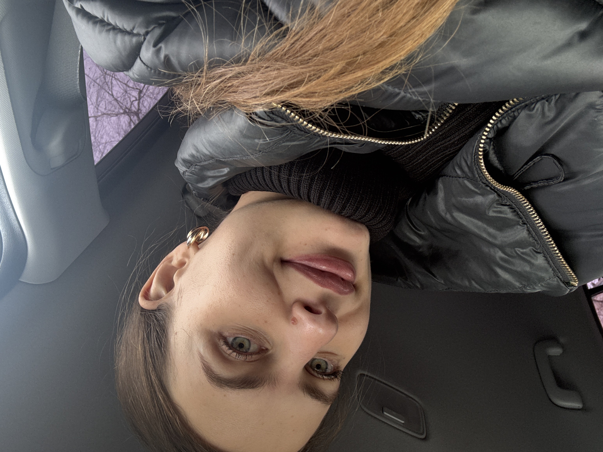 A young adult female is depicted in a close-up portrait, positioned inside a vehicle, likely a car, in Chaikovsky, Russia. Her head is tilted slightly to her right, and she is looking directly at the camera with a faint, closed-mouth smile. She has light skin, light green or hazel eyes, and brown hair with reddish-brown highlights, some strands of which are visible across her black puffer jacket. A small, light brown mole or blemish is present on the bridge of her nose. Her lips are a muted pink or plum shade. She wears a black ribbed turtleneck sweater beneath a black puffer jacket, which has a gold-toned zipper partly visible. A gold-toned double-hoop earring is visible on her right earlobe. The background consists of the car's gray headliner, a gray grab handle, and a side window revealing blurred, bare tree branches against a light purple-tinted sky, indicative of an outdoor environment during an overcast or colder period.
