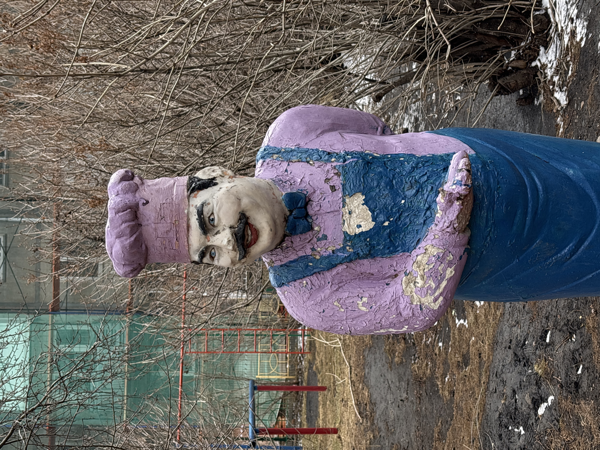 The image shows a weathered, painted concrete sculpture of a chef. The sculpture is positioned outdoors, appearing to be in a park or garden setting. The chef figure wears a lilac chef's hat and a lilac apron with blue details. The paint is peeling and chipped, revealing the concrete underneath. The chef's face features exaggerated features with a painted mustache and a smile. In the background, there are bare tree branches, a building under construction covered with green mesh, and a partially visible playground structure. The ground around the statue is covered with dry grass and some mud.