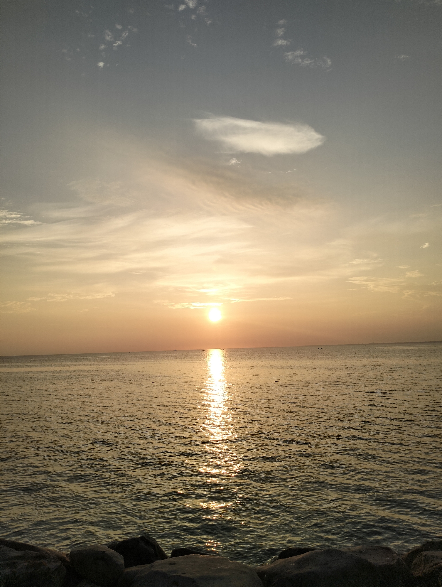 The image depicts a sunset over the sea, likely taken near Lhokseumawe, Indonesia. The sun is positioned low on the horizon, casting a golden reflection on the water's surface. There are some clouds in the sky and in the distance, there are several boats in the water. In the foreground are several large rocks. The overall scene conveys a sense of tranquility.